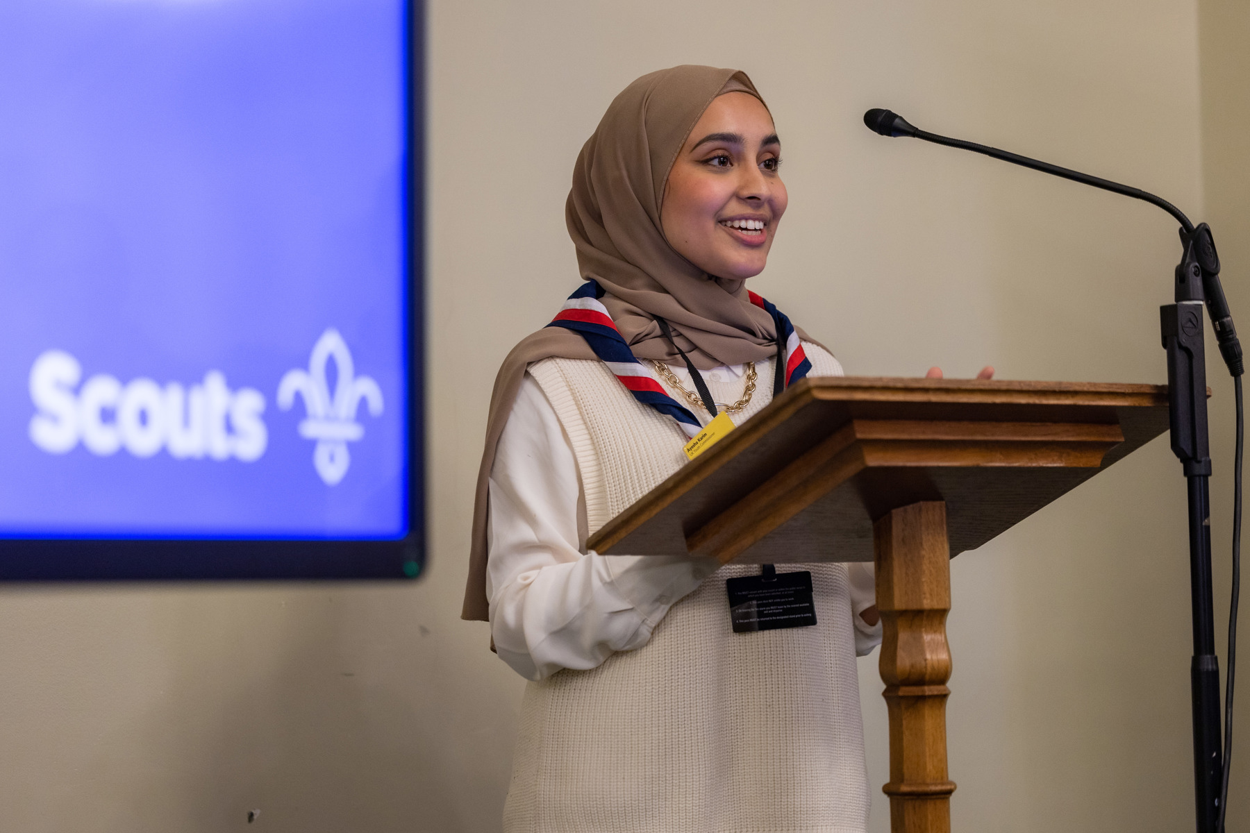 UK Youth Commissioner Ayesha Karim is standing up to speak in Parliament. She's wearing a red white and navy necker and stands in front of a microphone. Behind her is a purple screen with a white Scouts logo on.