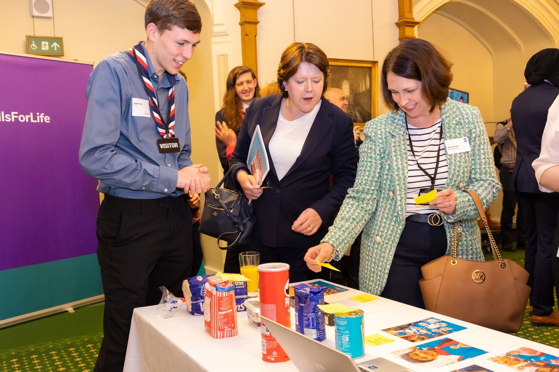 Three Scout members at Parliament are looking at a table with crisps, flour, bread, rice and beans and leaflets. One visitor in a green and white blazer jacket holds up some yellow sticky notes and each member is looking at them to read what they say.