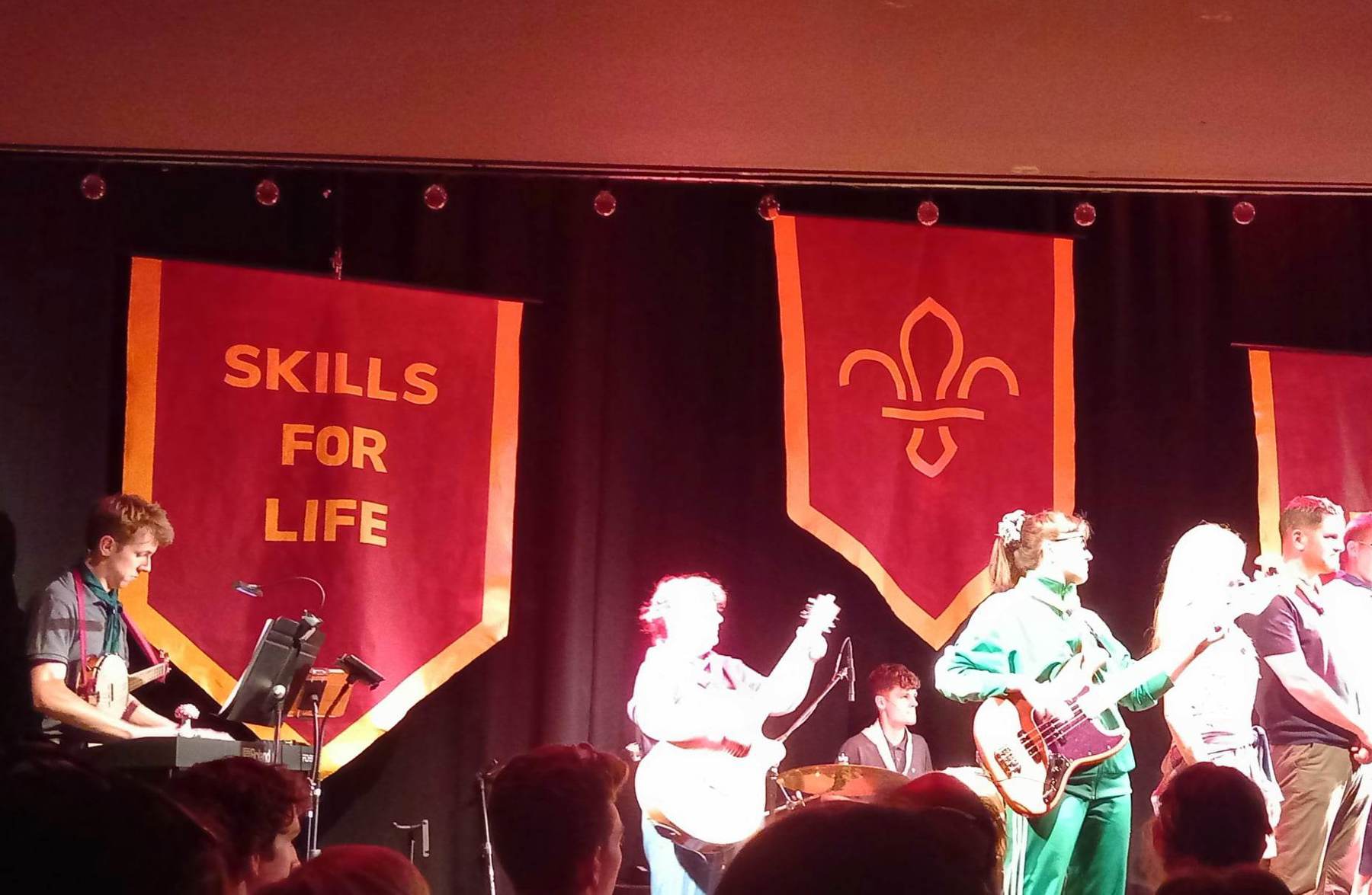 Five musicians are stood on a stage with banners behind their heads. One banner says Skills for Life and the other has a fleur-de-lis.