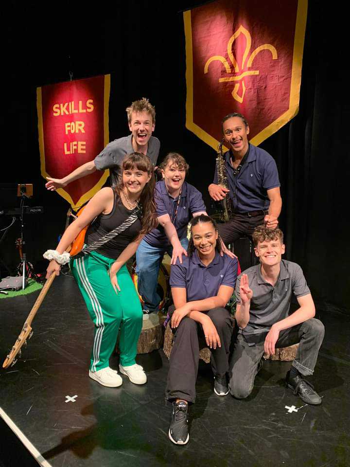 Six cast members of Scouts! The Musical are stood crouching down on the stage in their costumes smiling at the camera.