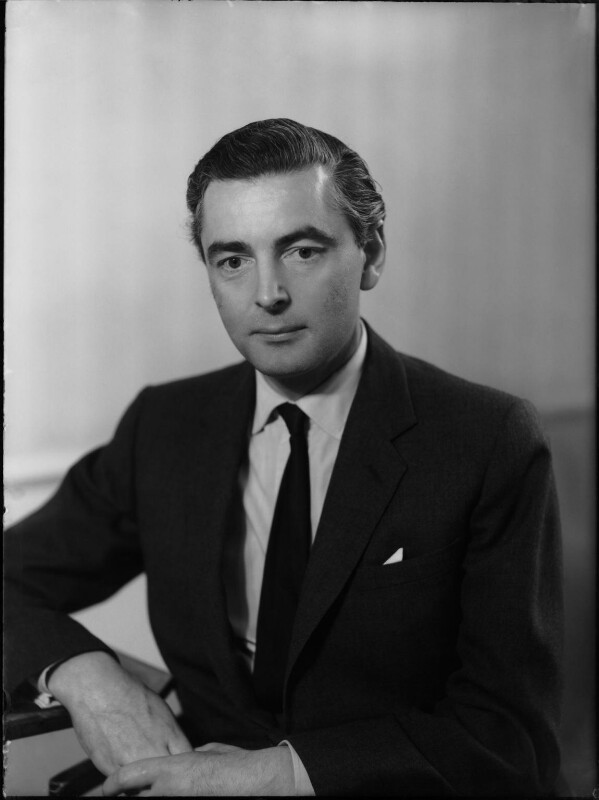 The 13th Earl Of Airlie with dark slicked back hair and dark suit leaning on a desk in 1961