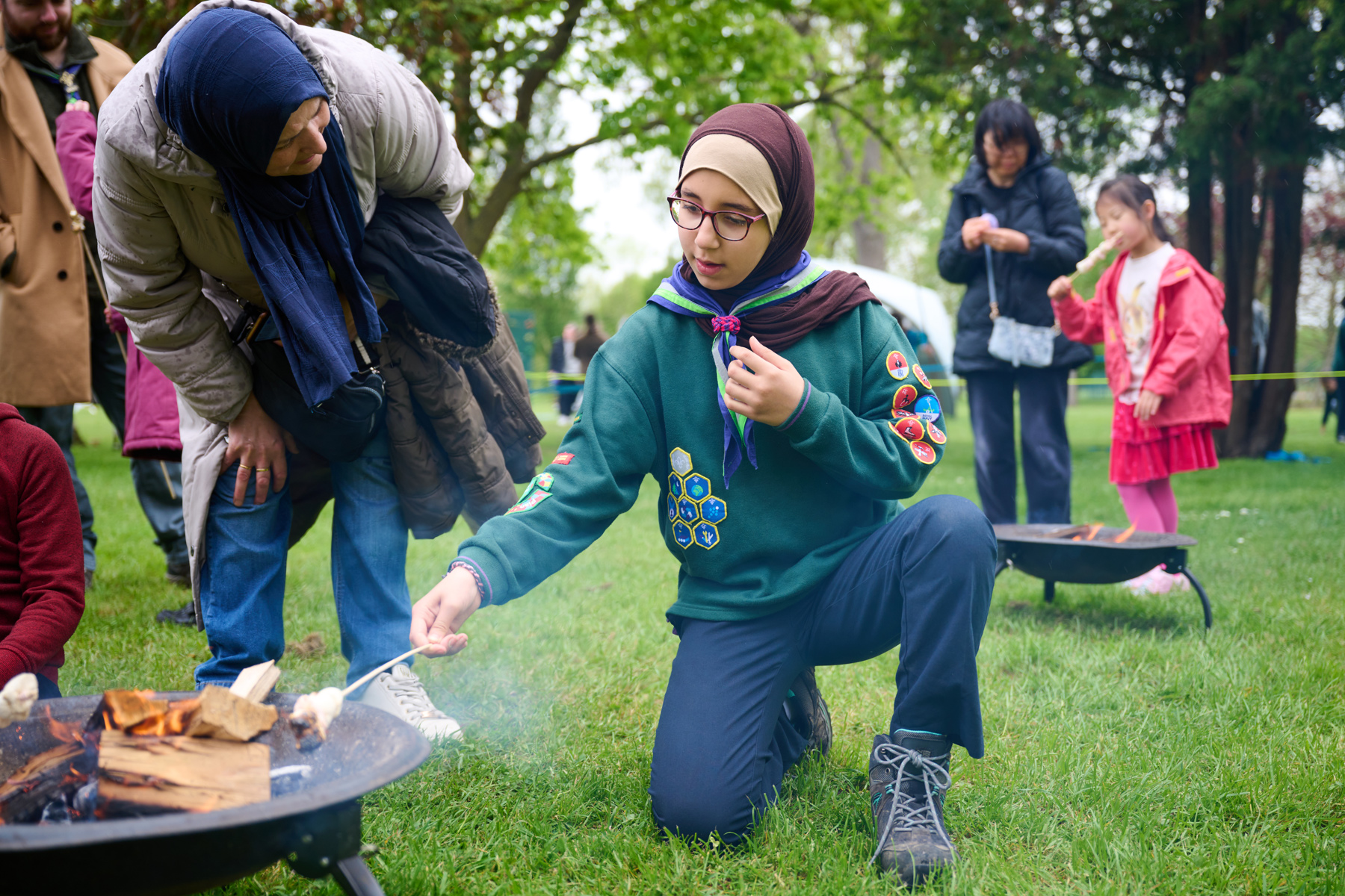 A Cub kneels by a campfire with a stick as a volunteer stands next to them.