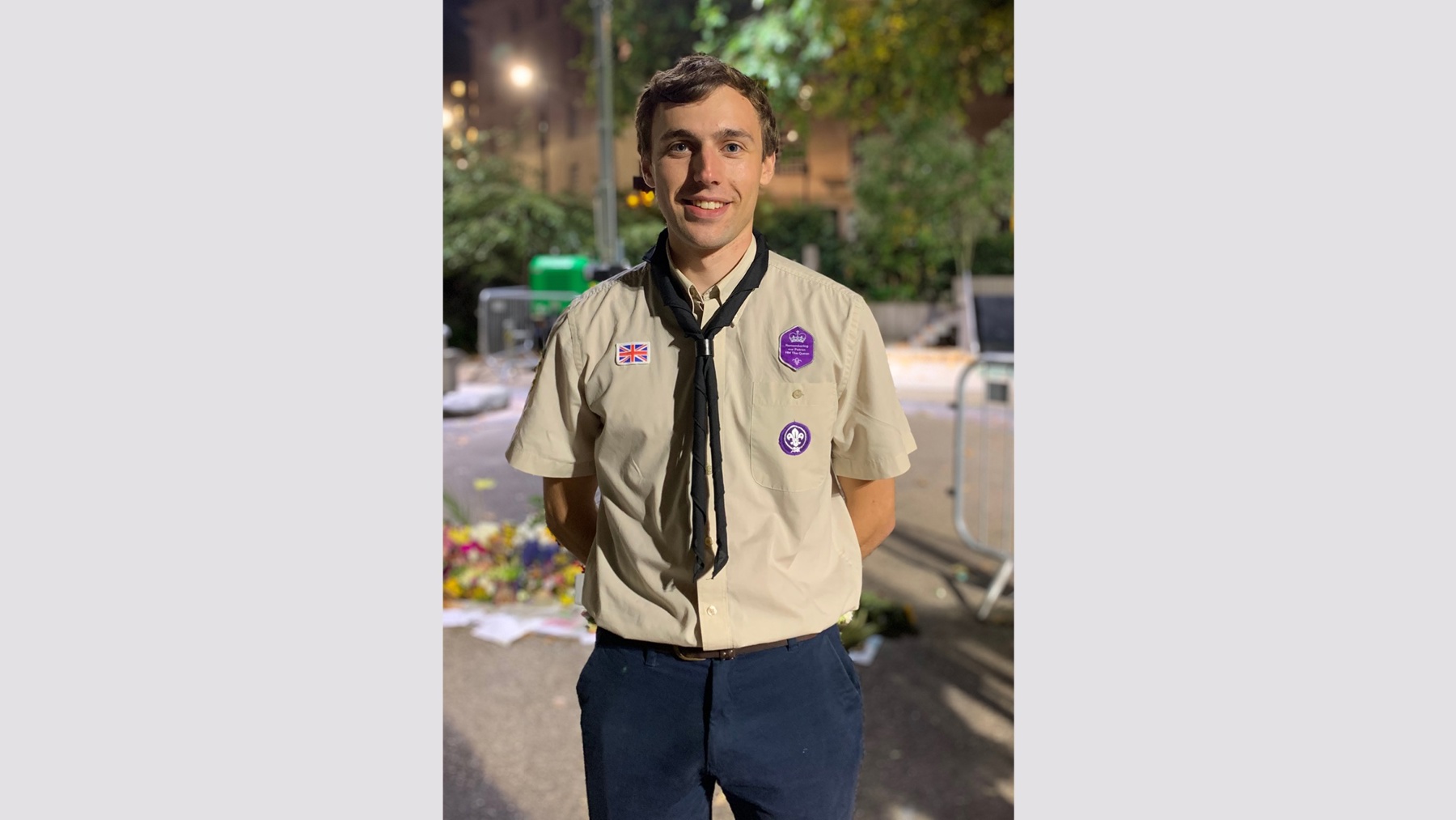 Aaron is standing with his hands behind his back and is wearing his Scouts uniform. He's wearing a beige shirt with badges on and a black necker. He's stood outside at night and there's a streetlight on in the distance. Behind him blurred in the background are flowers and a metal railing.