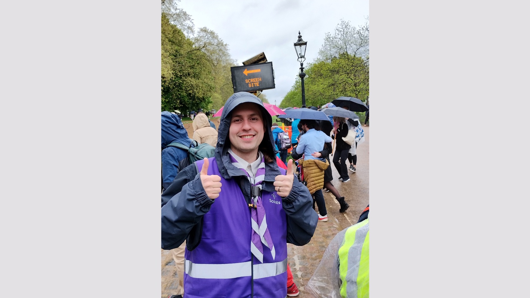 Luke's holding his thumbs up to the camera and smiling. He's got a purple hi-vis vest on with the Scouts logo in white, along with a purple and white necker. He's wearing a rain jacket with his hood up, and he's outside near some trees. There are people behind him holding umbrellas up and wearing rain jackets too.