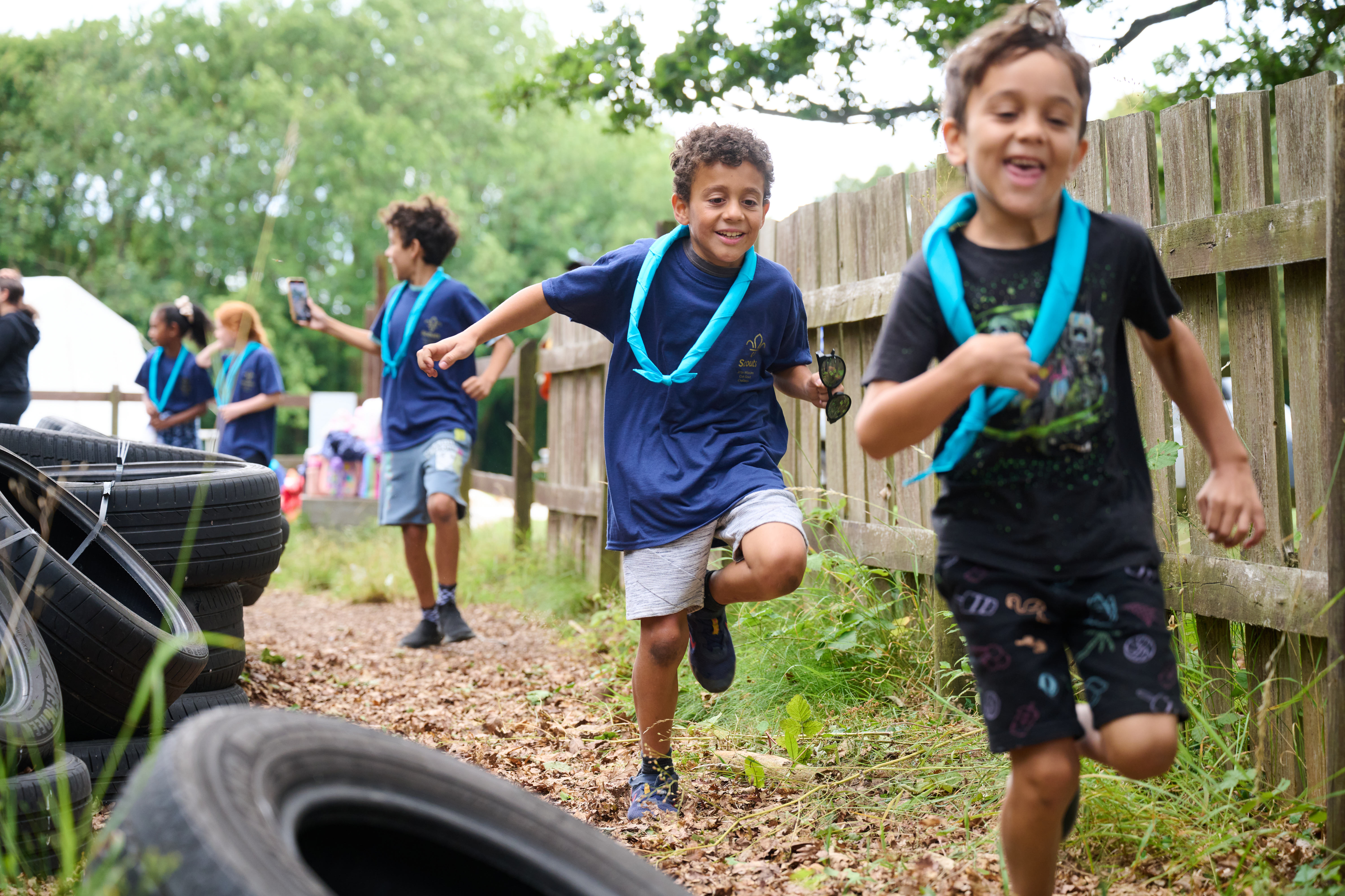 Two Cub Scouts are wearing light blue neckers and running outside with a brown fence to their left. On their right are old tyres on the ground, and they're both wearing t-shirts and shorts. They're looking at the ground and smiling as they run.