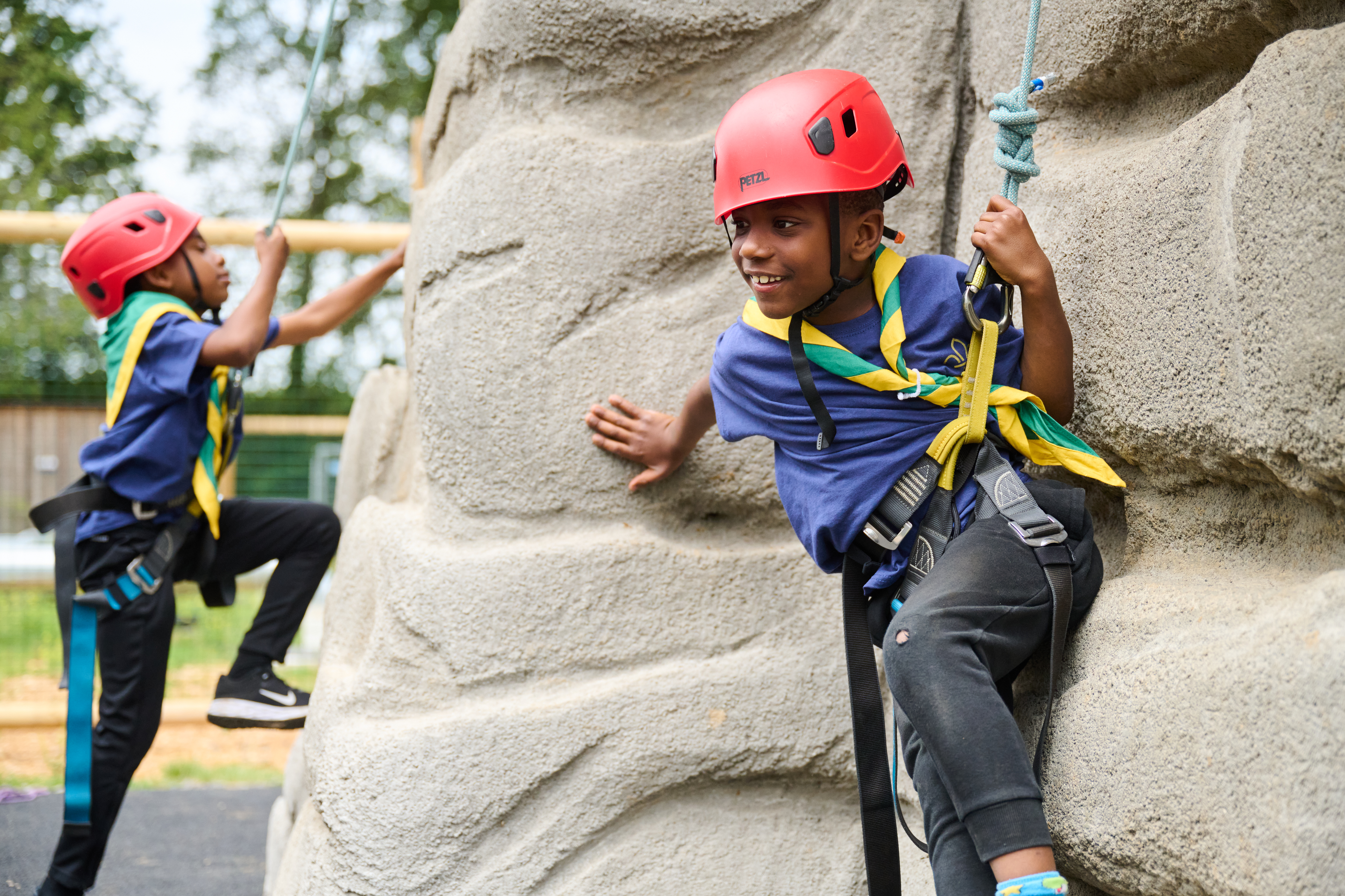 Two Scouts are wearing helmets, t-shirts and neckers while attached to rope near a climbing rock. The Scout to the left has their left leg on the rock and is about to start climbing up, and the Scout to the right is leaning on the rock with their left hand against the rock to support them.