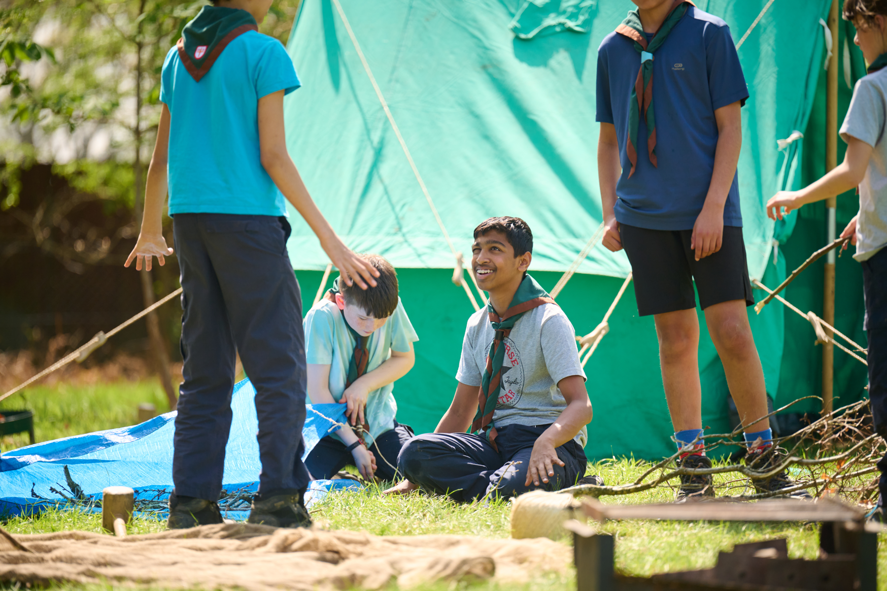 Four Scouts stand and sit on a campsite while pitching up.
