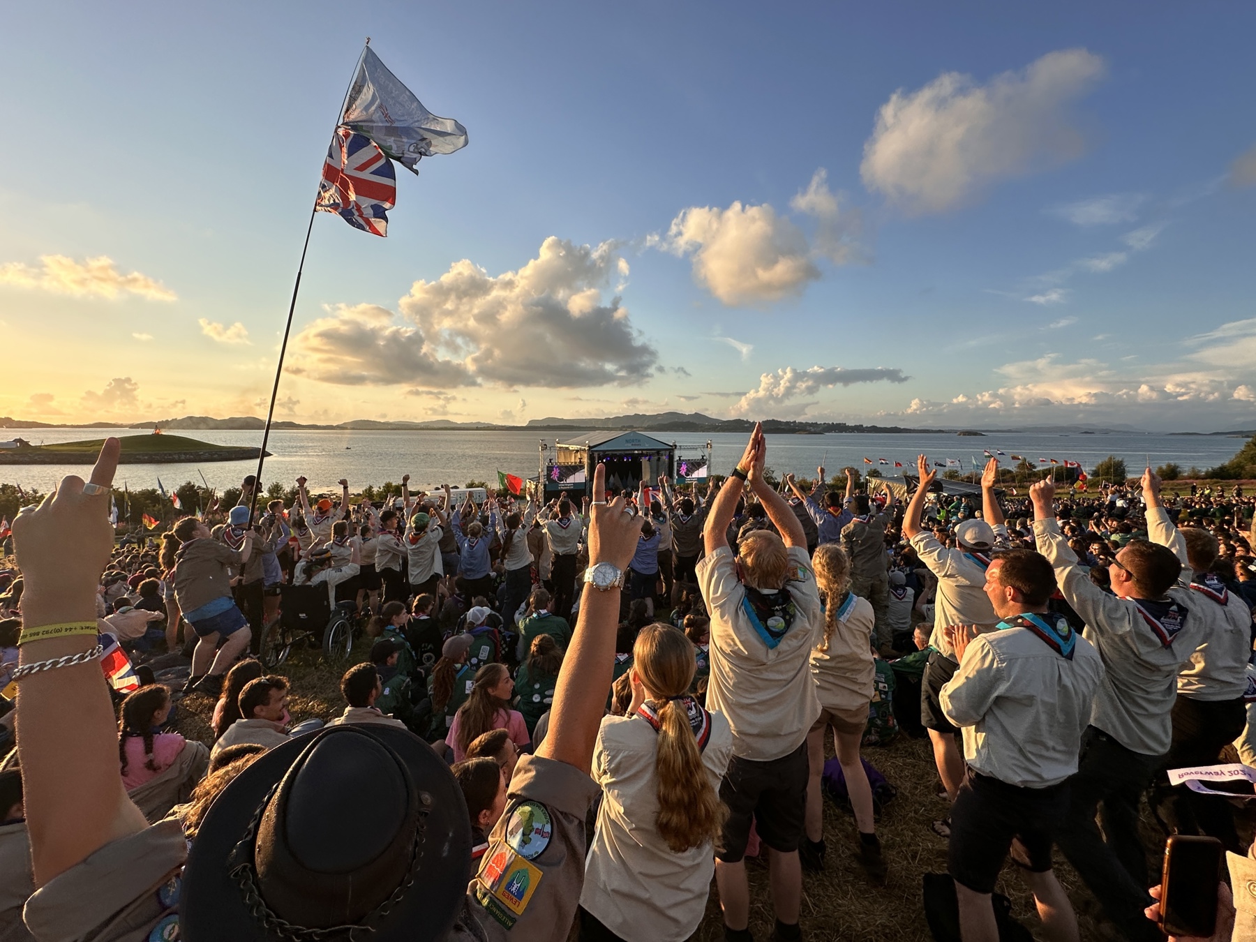 A very large crowd of Scouts are gathered facing the Roverway stage which is placed in front of the lake while the sun is setting. The Scouts wearing uniform are stood with their hands in the air and cheering, while someone waves a Union Jack flag on a pole.