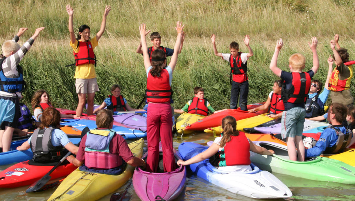 A group of Scouts wearing life jackets stand up in kayaks reaching their arms into the air.