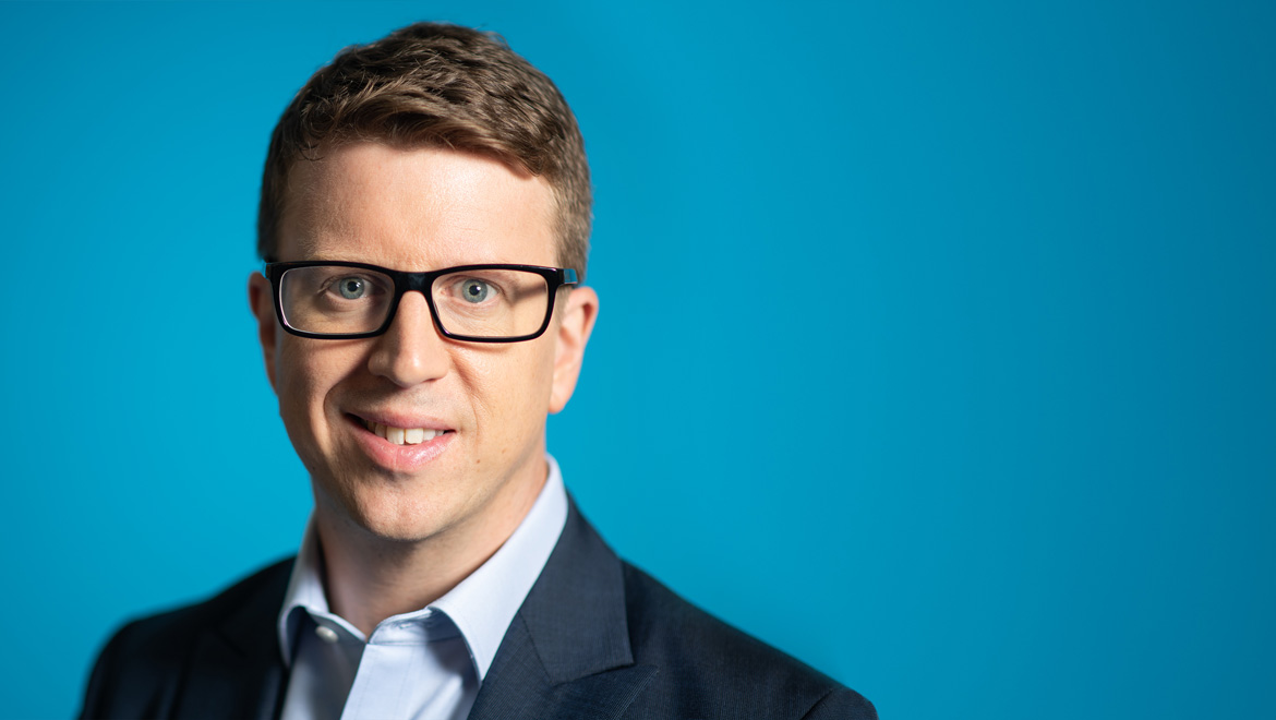 Headshot of Matt Hyde smiling at the camera wearing glasses, a light blue shirt and navy suit jacket