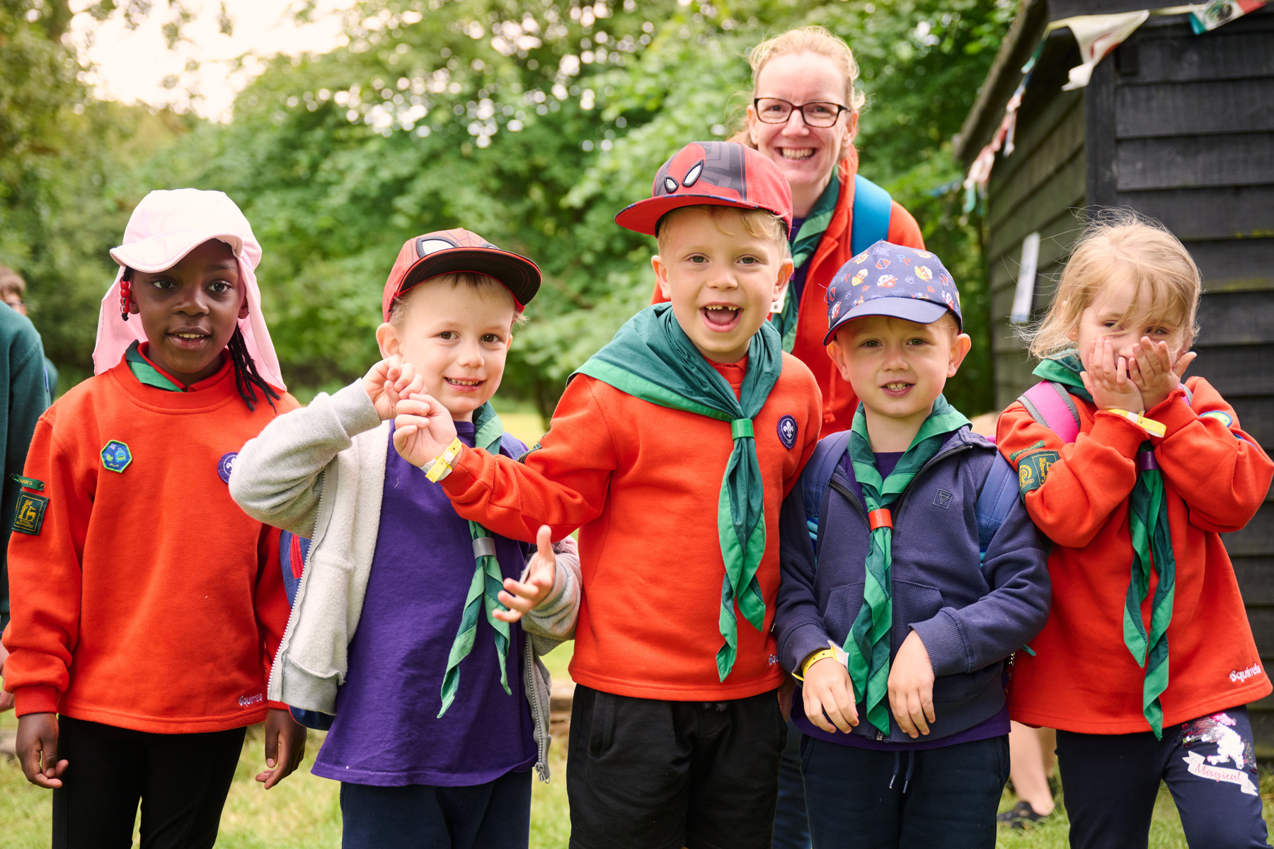 The image shows Squirrels in their uniform smiling at the camera. Thee are 5 and they are stood in a line. Just behind them is a volunteer and they're stood in front of trees with a black wooden building to their right.