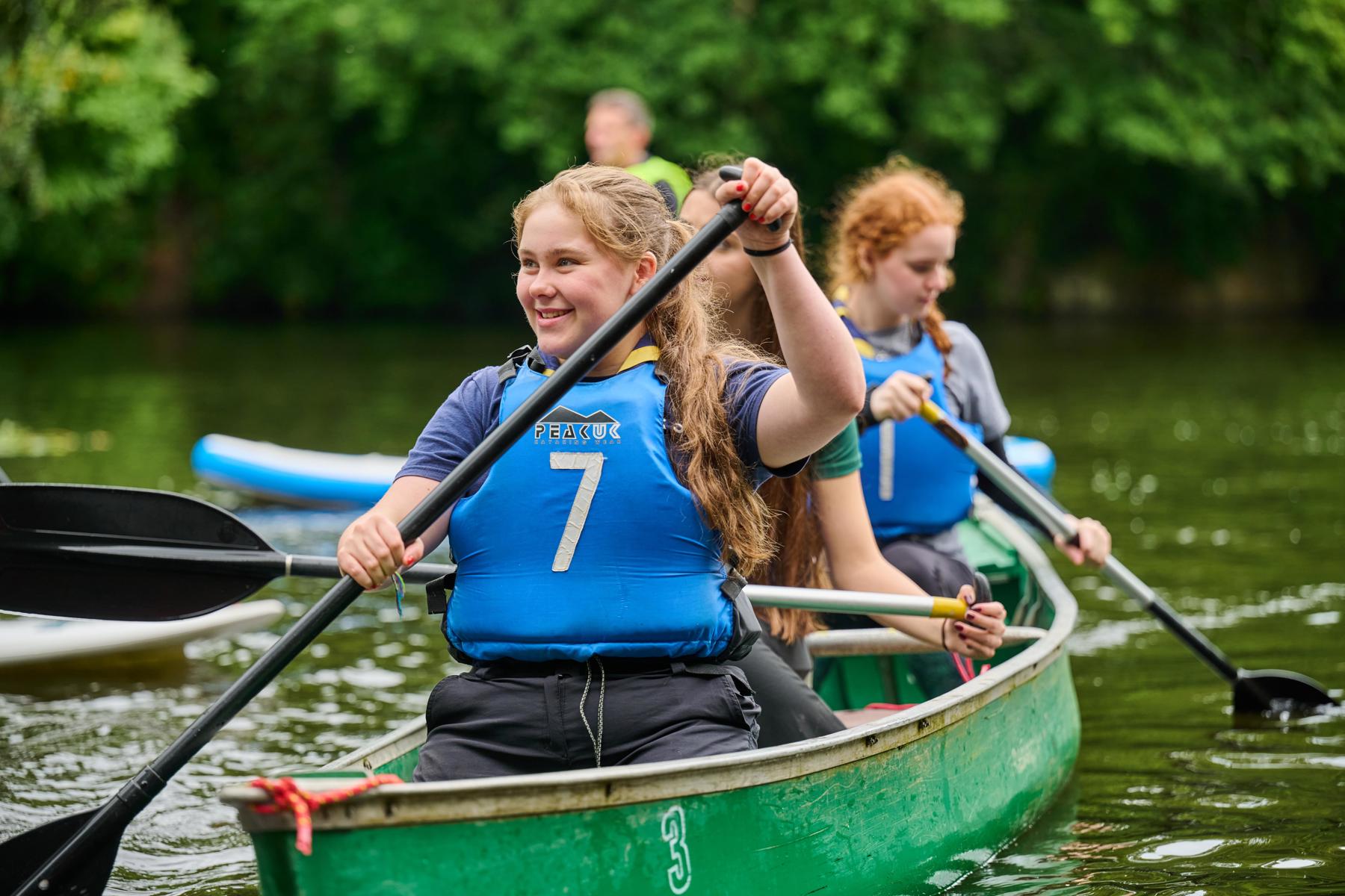 Three smiling Explorers in blue lifejackets paddle a green canoe on a lake.