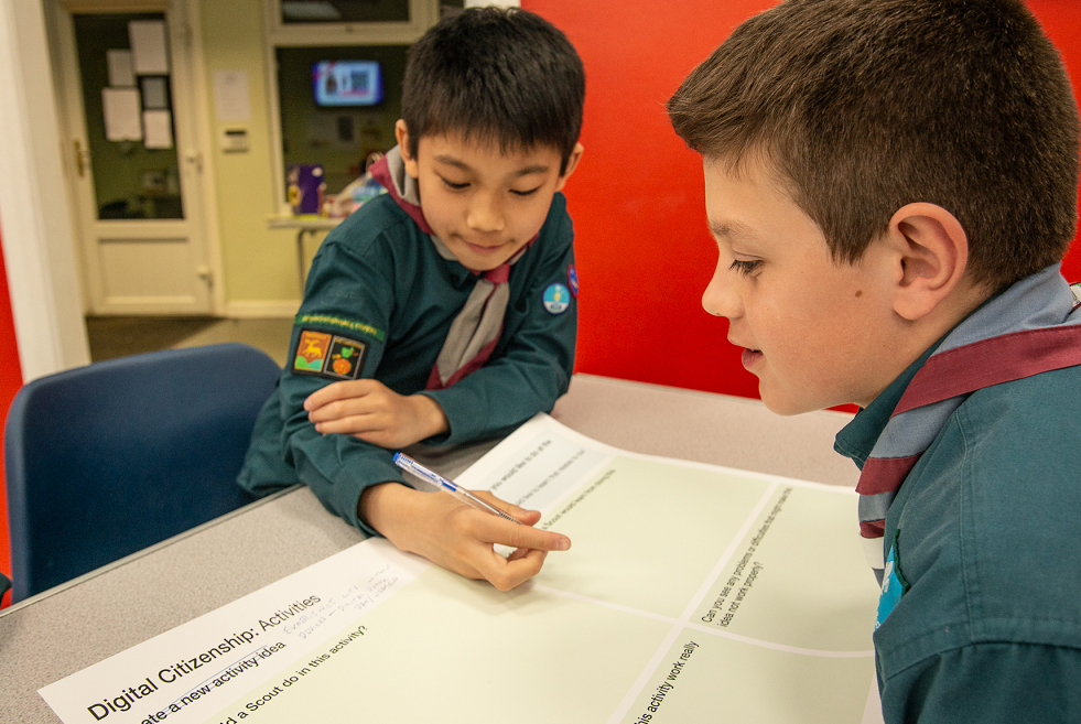 Two scouts around a table looking at a piece of paper with 'Digital Citizenship Activities' written at top. Scout on the left hand side is holding a pen. 