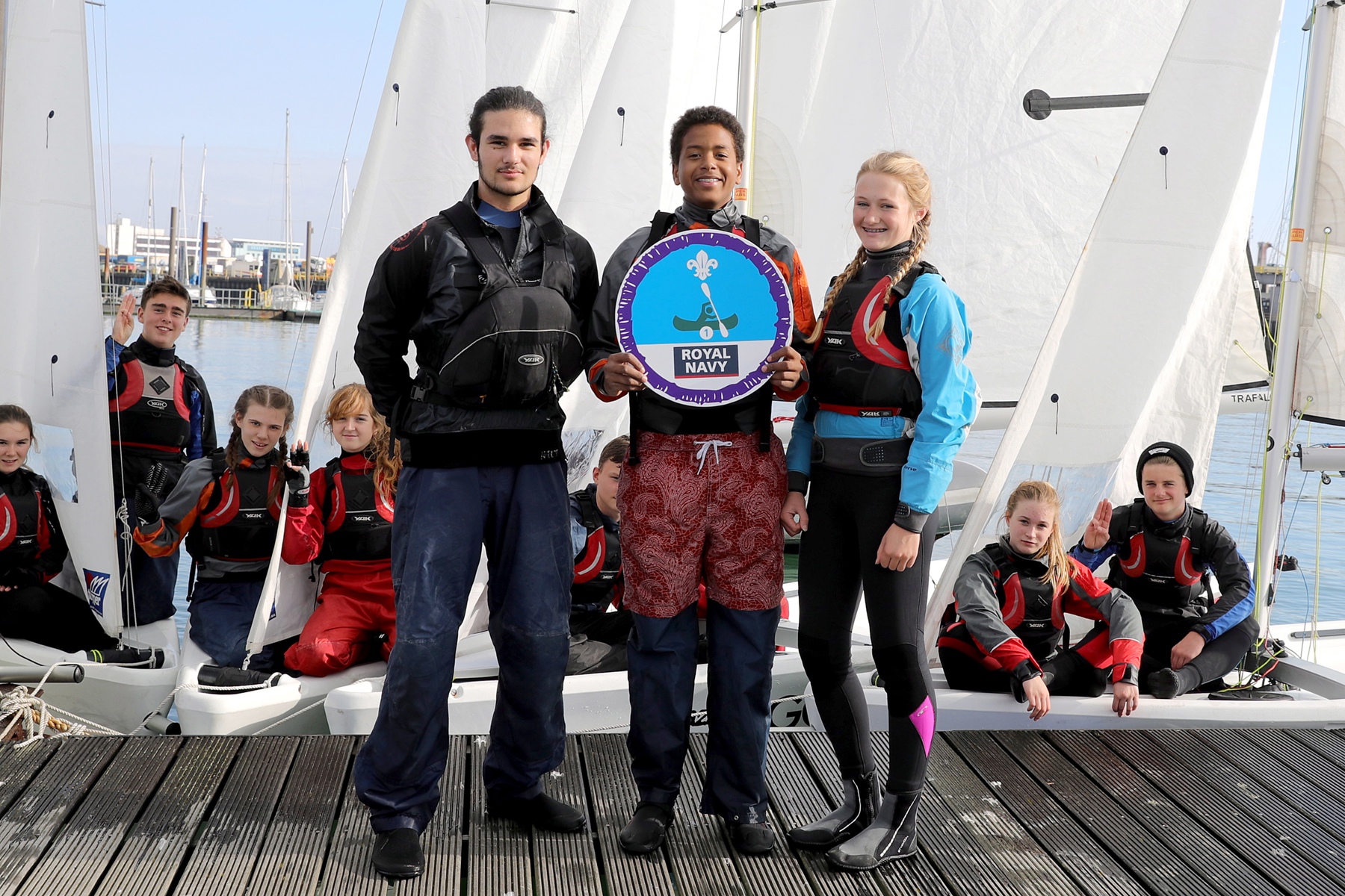 Three young people holding up a giant Time On Water badge who have just been awarded their badge, standing in front of a moored sail boat