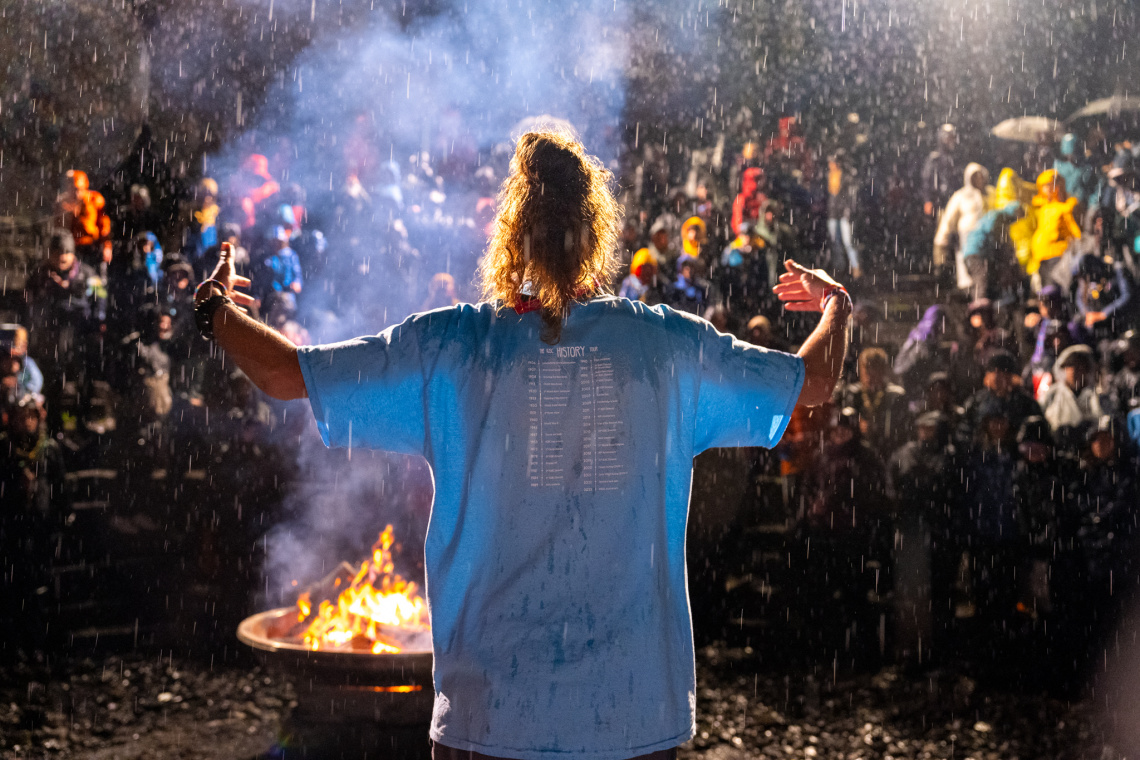The image shows a Scout in a light blue t-shirt with their back to the camera. They're holding their arms out to the sides and they're a distance away from a blazing campfire in front of them. A distance behind the campfire are lots of other Scouts gathered together. You can see the rain pouring down.