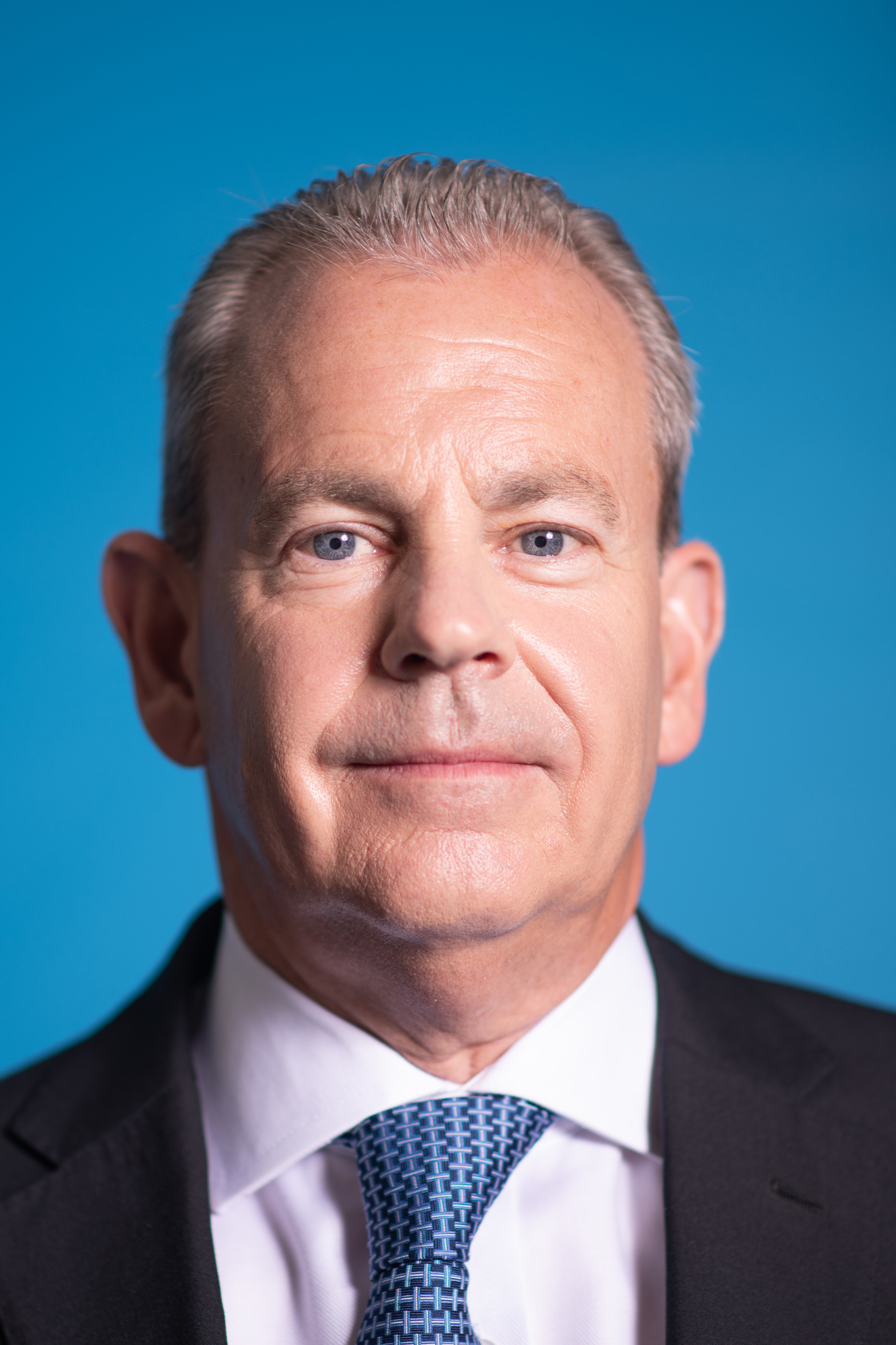 Headshot of Mark Hislop looking at the camera wearing a white shirt, blue patterned tie and black suit jacket
