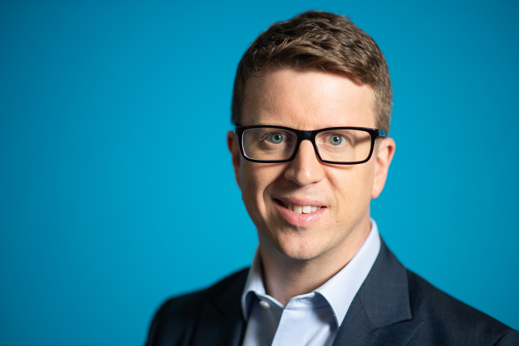 Headshot of Matt Hyde smiling at the camera wearing glasses, a light blue shirt and navy suit jacket