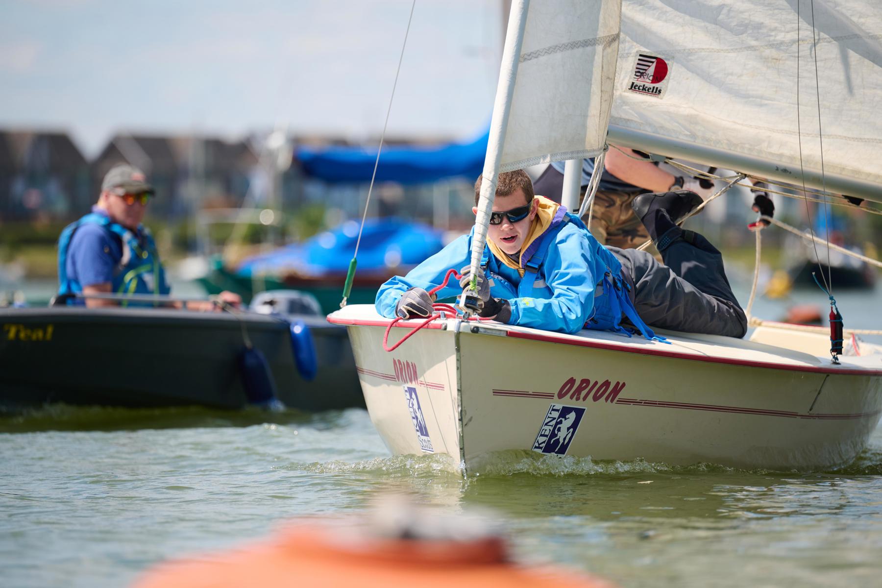 A Scout in a blue jacket and lifejacket lays on the front of a sailboat in the water.