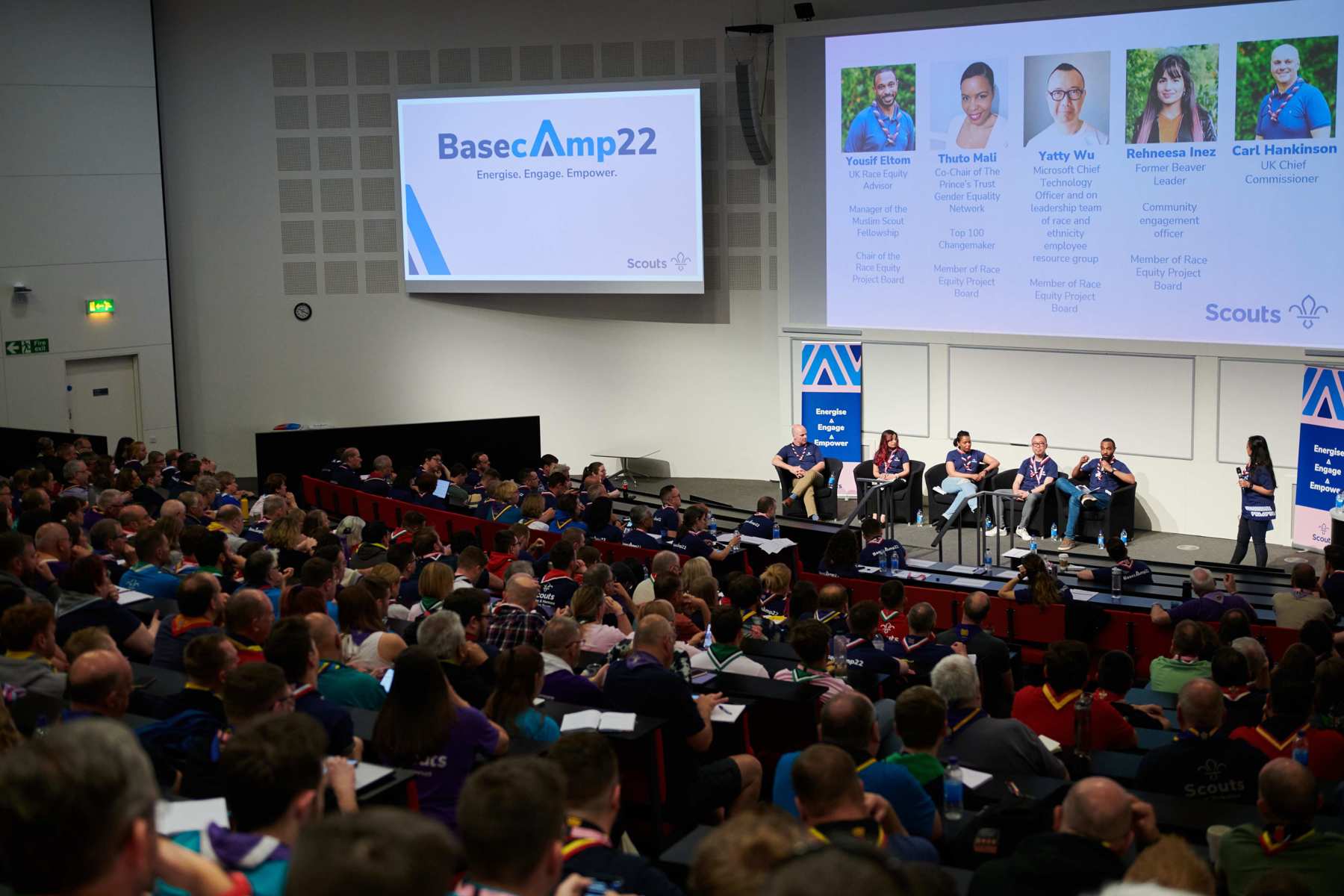 The image shows a crowd looking at a screen and stage at Basecamp 22. There are presentations on the screens with 4 people sat on the stage giving a talk.