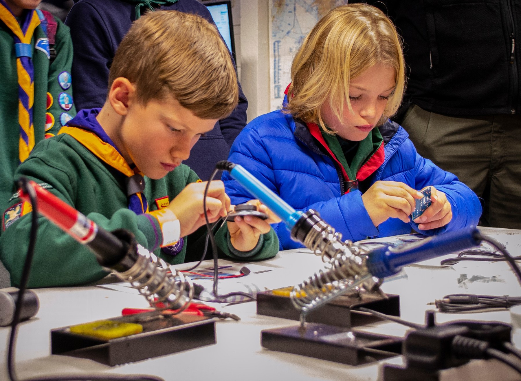 Two Scouts, one boy and one girl, are sat at a table building an electrical circuit. They are concentrating on the circuit boards and are sat with soldering irons.
