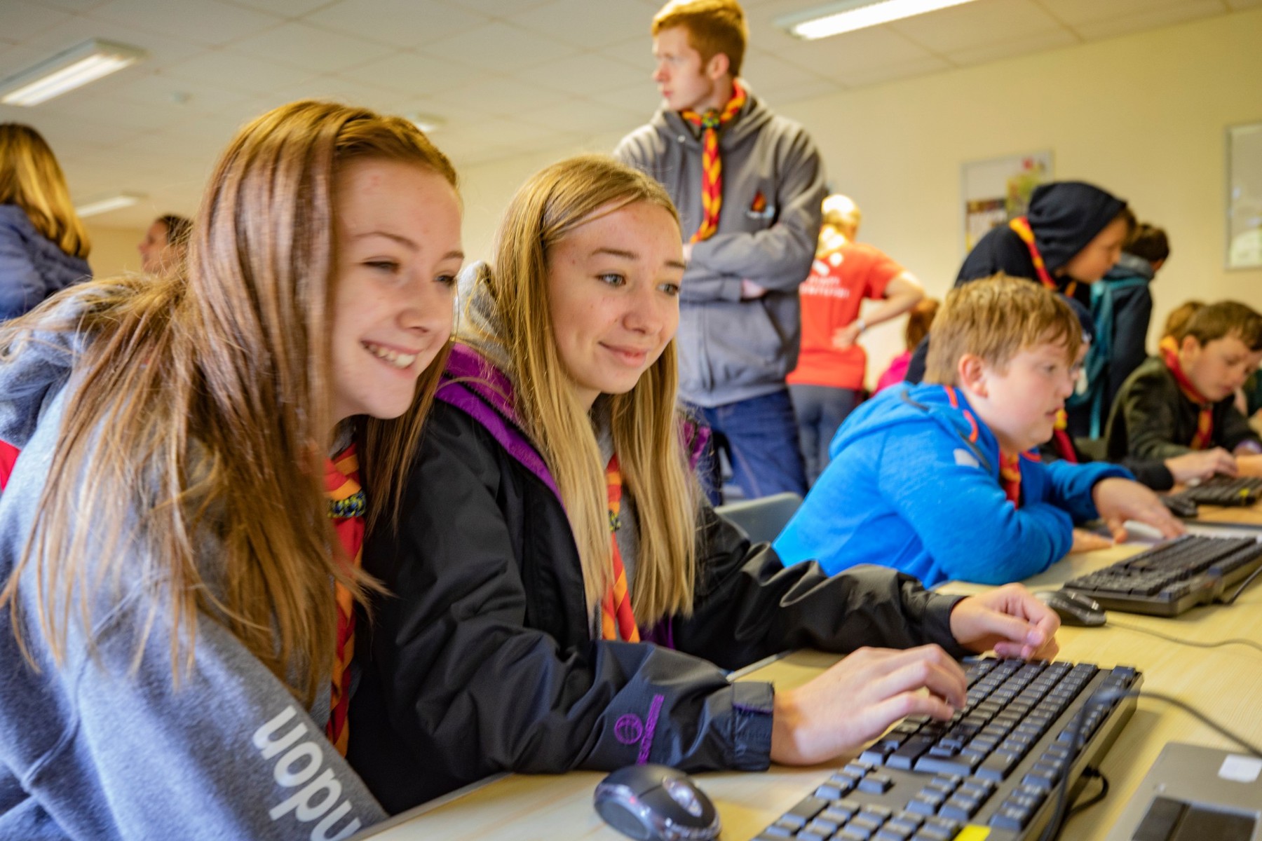 A group of Scouts are in a room, sat in front of computers. Two girls are in the front of the image, wearing neckers and using a keyboard. They are smiling and engaged.