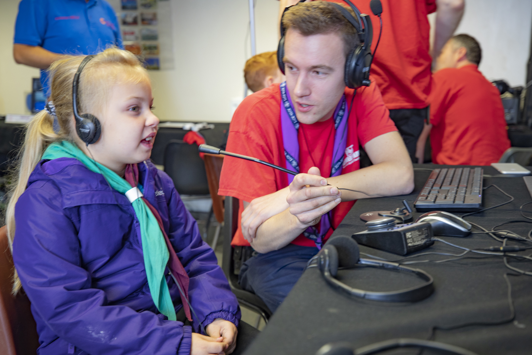 A young girl is taking part in JOTA-JOTI with an adult helping her. She is sat down, wearing headphones and speaking into the microphone by the desk. The adult is helping to hold the microphone.