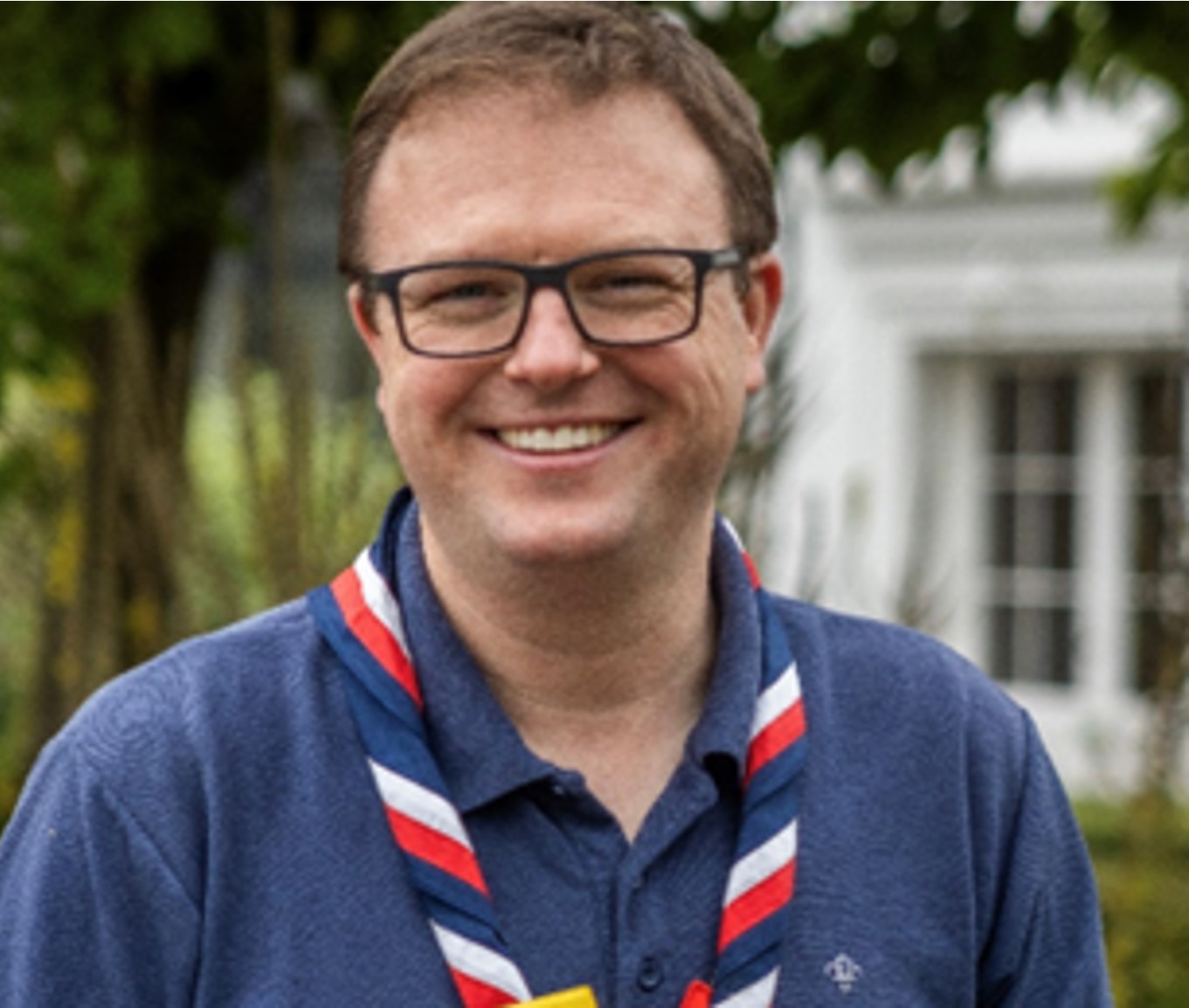 Rob is smiling and wearing black glasses, a blue Scouts polo and a red, white and blue necker.