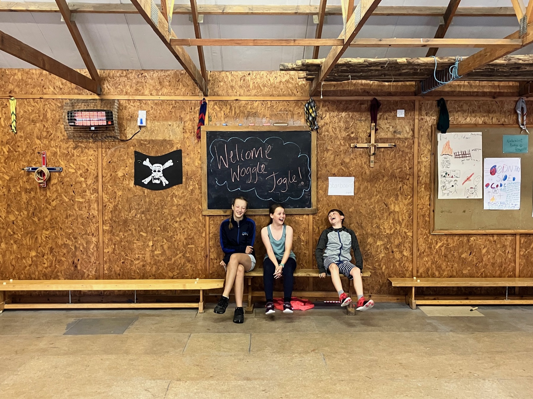 The image shows Bethan, Hattie and Ollie sat on a bench inside a wooden hut. On the blackboard behind their heads, 'Welcome Woggle JOGLE' is written. They're all laughing together. 
