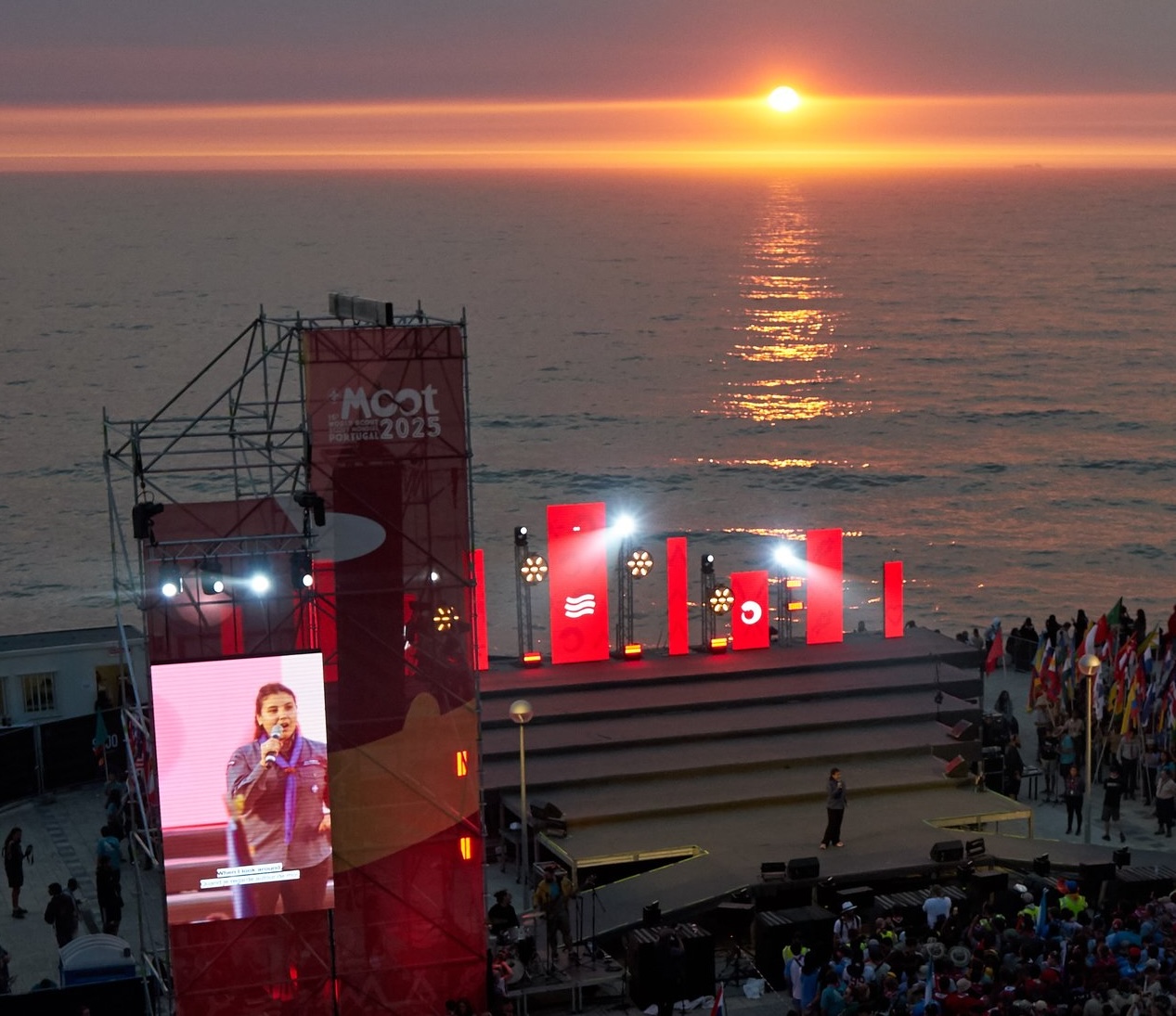 The image shows the sunset at the 16th World Scout Moot in Portugal. The sun is setting over the sea, and there are Scouts gathered in front of a stage just before the ocean.