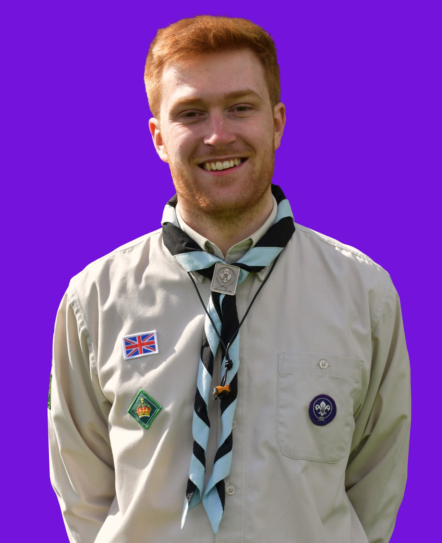 This image shows a man smiling at the camera wearing a Scouts shirt uniform and a blue striped necker. He has ginger hair and a ginger beard and he stands with his arms placed behind his back in front of a plain purple background.