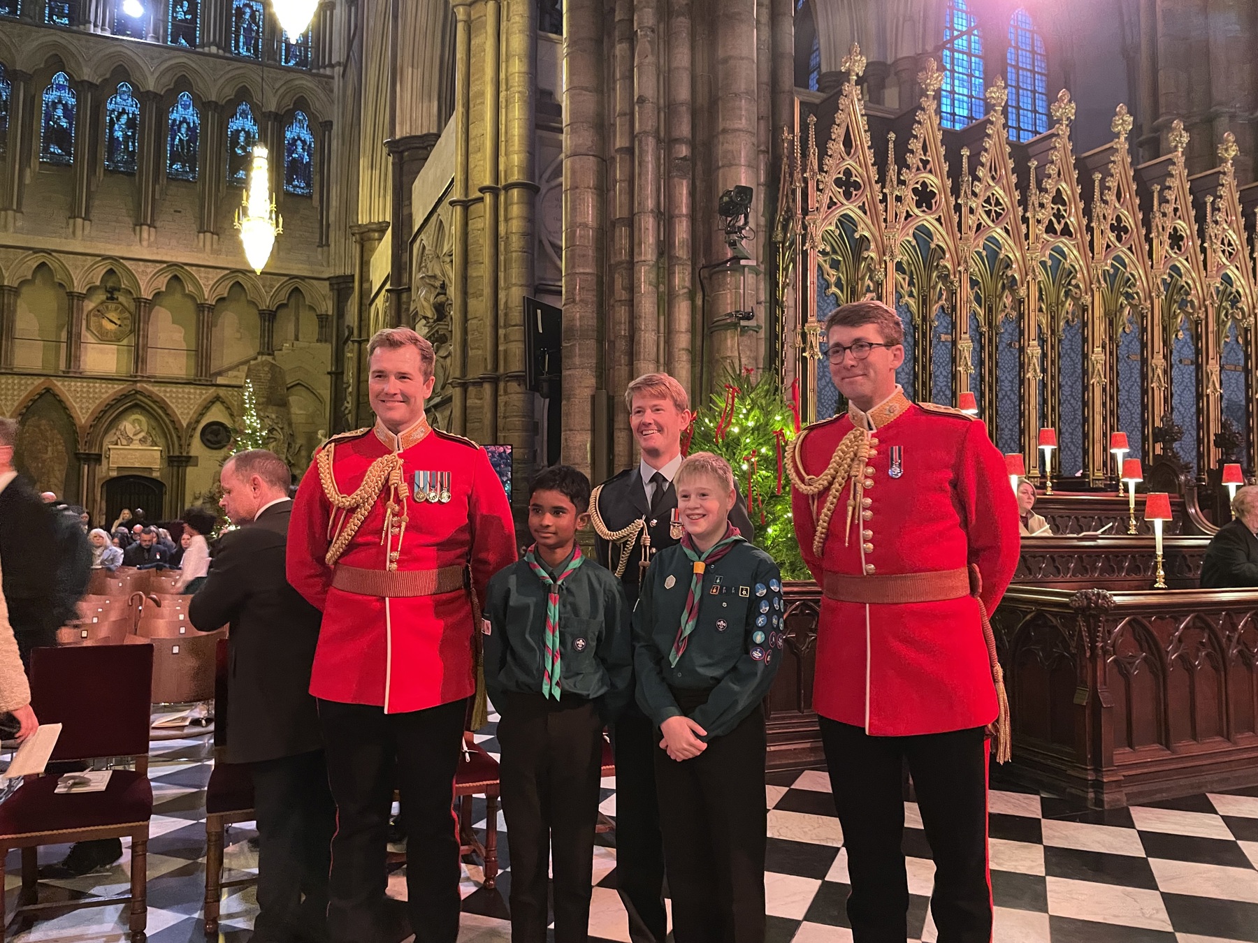 The image shows two Scouts in uniform and neckers looking past the camera. They’re stood with three men in uniform with their hands behind their backs. They’re all stood inside Westminster Abbey and there’s a Christmas tree behind them all.