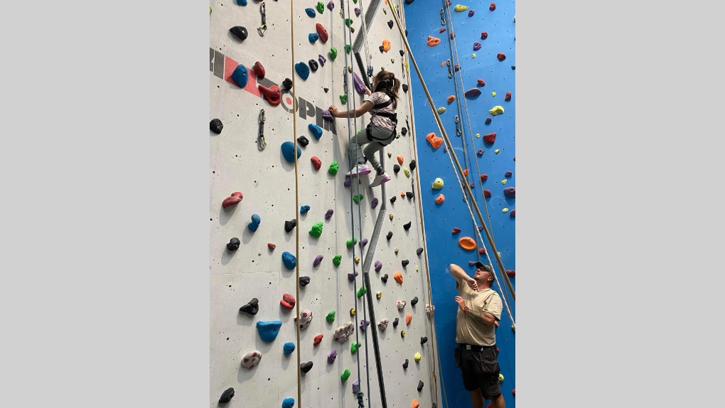 The image shows Sophie climbing up a climbing wall. She's holding onto and facing the wall and wearing a safety harness. There's a volunteer on the ground who's looking up at Sophie to support her.