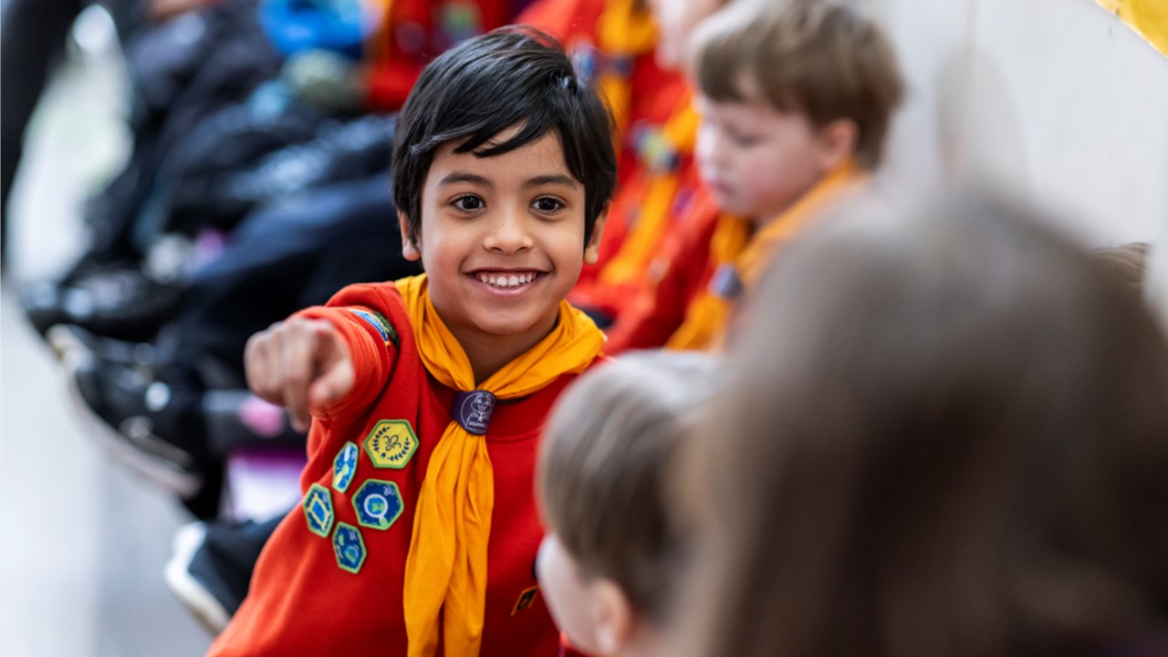 A Squirrel in a red jumper and orange necker points while smiling.