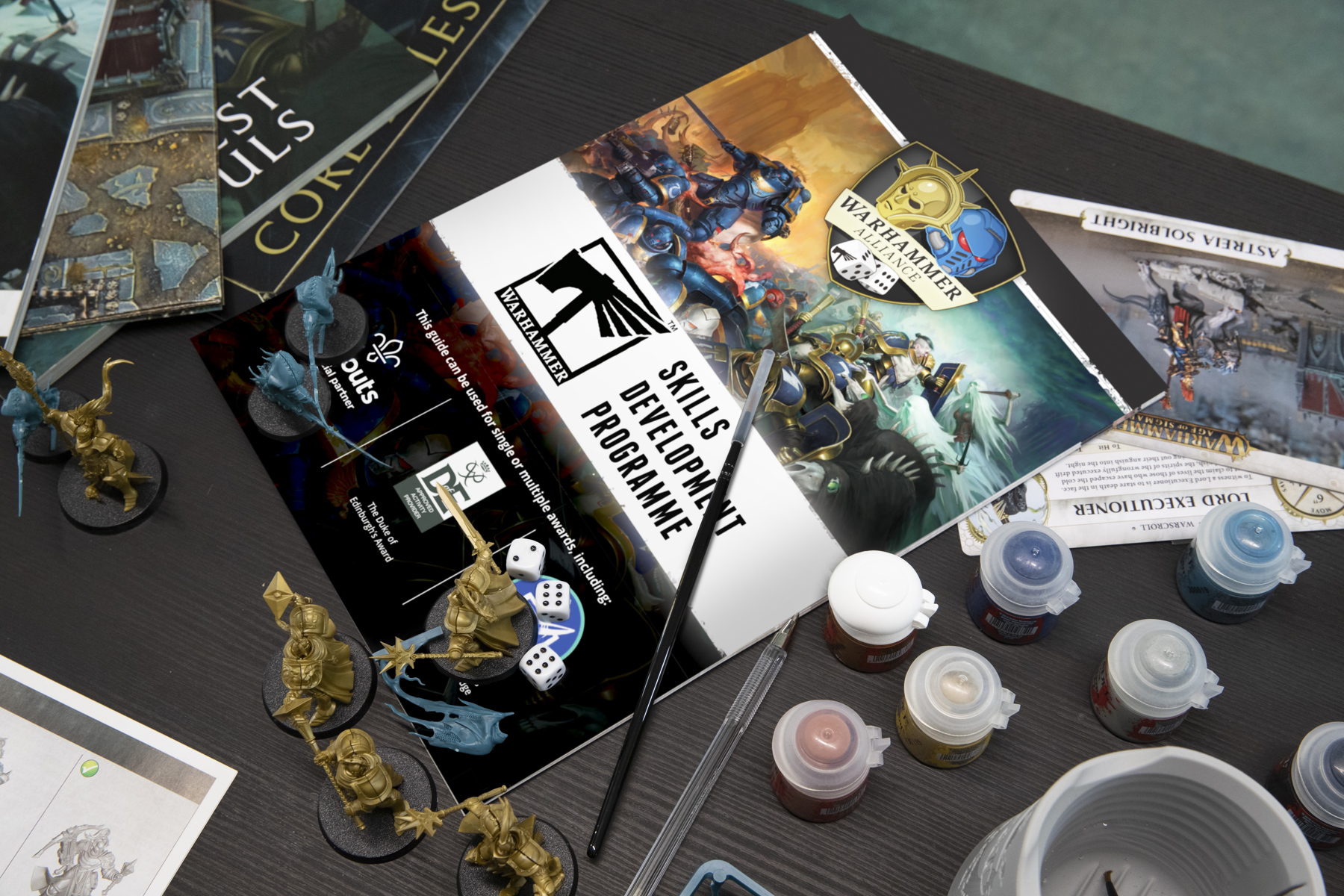 Warhammer Skills booklet on a table with Warhammer figurines, paint brushes and paint pots