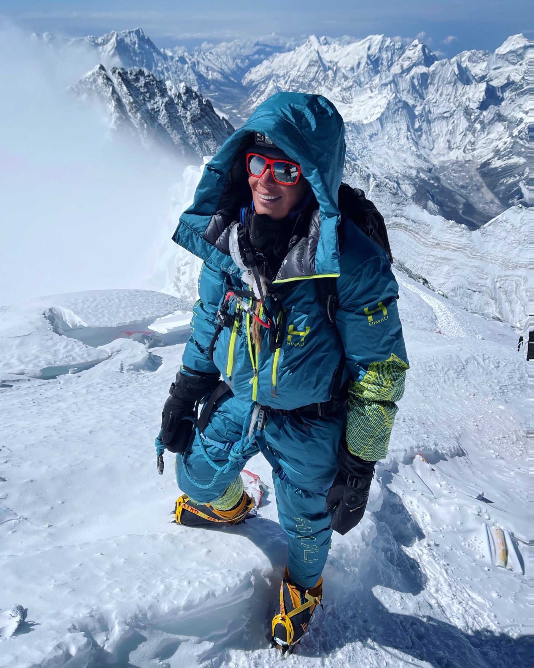 Kirsty Mack is stood on snow surrounded by mountains as she hikes up Everest. She's wearing goggles, gloves, blue trousers and a blue winter jacket. She smiles at the camera.