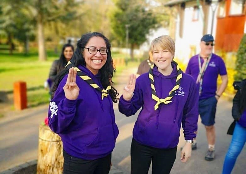 Two female Scouts in purple hoodies and yellow and black neckers are smiling at the camera. They are doing the Scout sign with their right hand. One has black hair and glasses, while the other has short blonde hair.