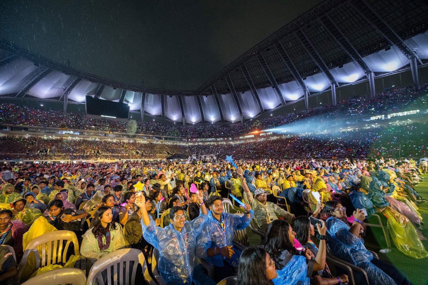The image shows a group of Scouts gathered in a crowd inside a stadium at a World Scout Jamboree. They're wearing blue and yellow rain ponchos and holding light up toys while sitting on plastic chairs.