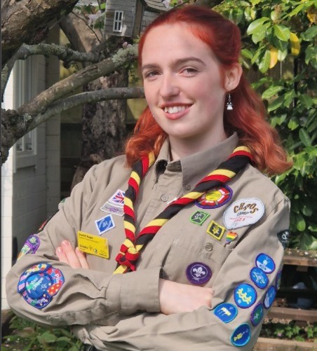 Scarlett, with their arms folded, smiles at the camera while wearing a khaki shirt and yellow, red and black necker.