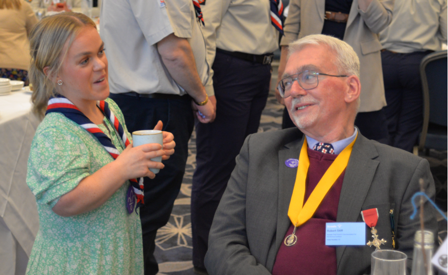 Bob is to the right of the image, sitting down and looking to his left with a medal around his neck. To his left is Scouts Ambassador Ellie Simmonds who's wearing a red, white and navy necker and holding a mug in her hands.