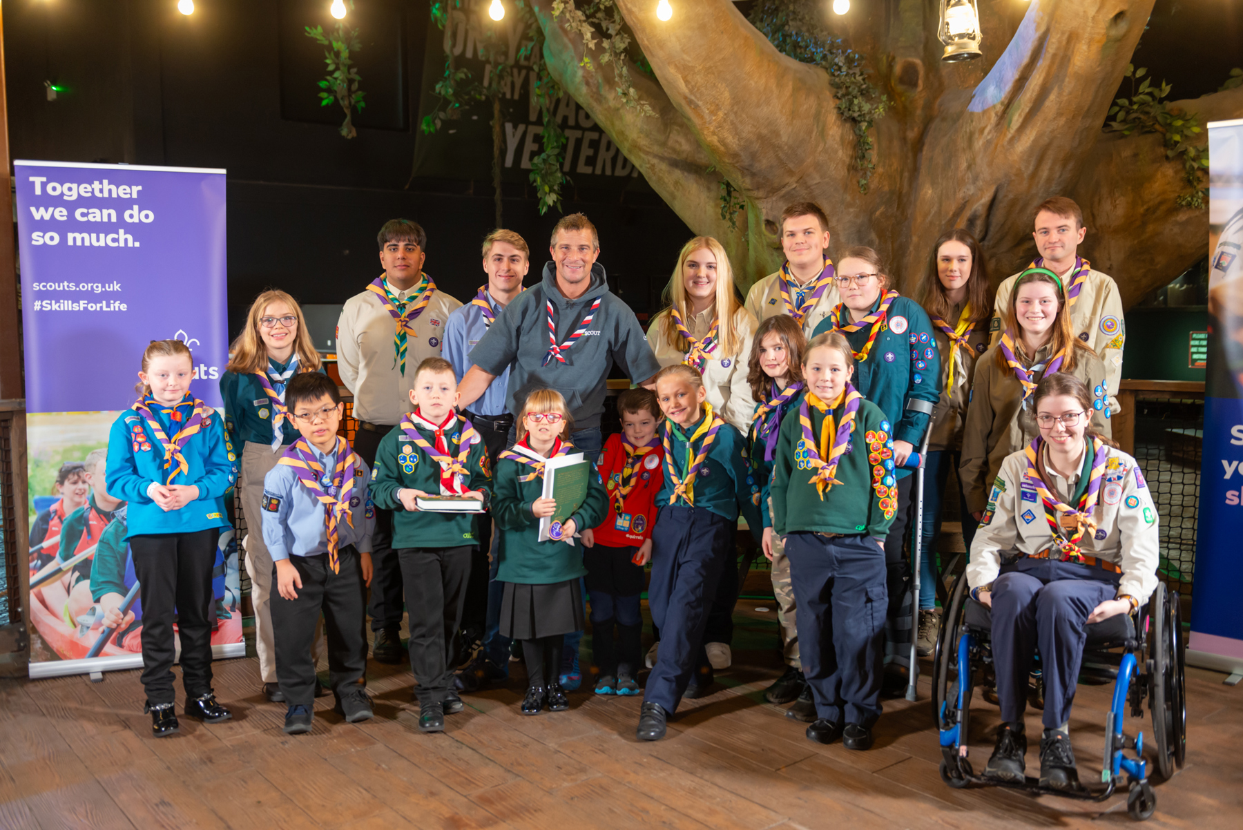 The image shows Chief Scout, Bear Grylls, stood with the 2023 Unsung Heroes. They're all in Scout uniform and pictured in front of an artificial tree with string lights above their heads. They're all smiling at the camera and to the left of them is a Scouts branded post, saying 'Together, we can do so much' and 'Skills for Life'. 