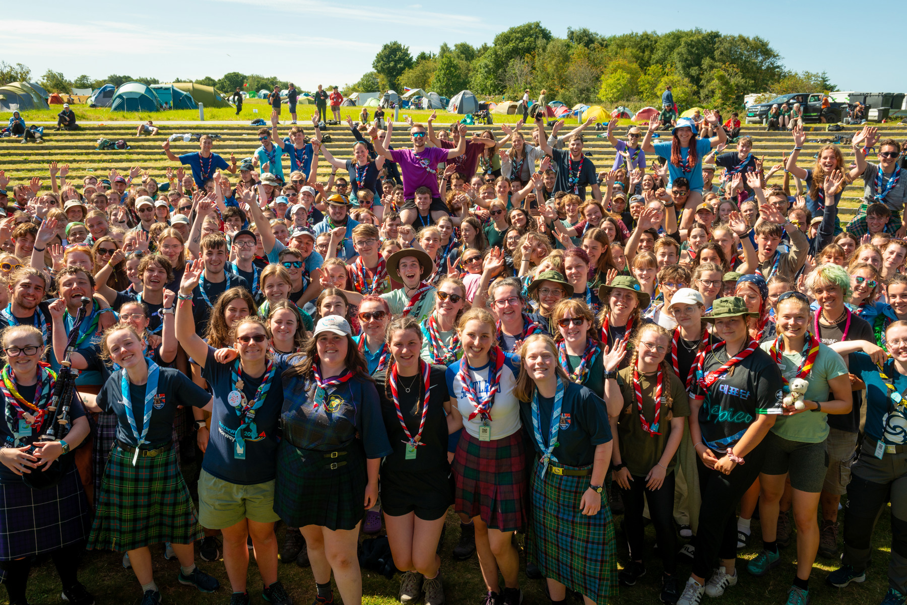 A large group of Roverway participants all stand together outdoors in a campsite smiling for the camera, some are wearing neckers and have their hands up, cheering.