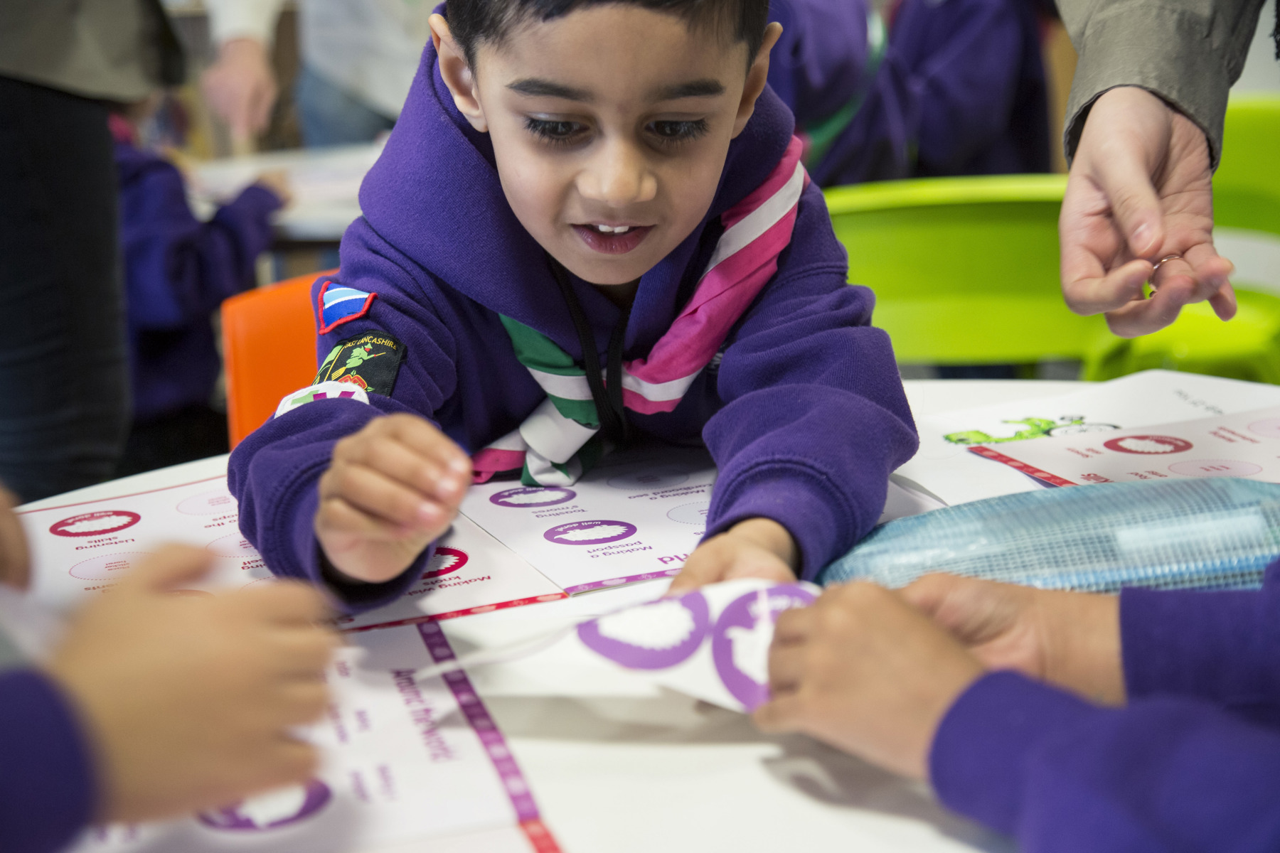 A young Scout boy is engaging in an activity, wearing a purple jumper and pink and green necker. He is leaning over the table while smiling to help put stickers onto a sheet.