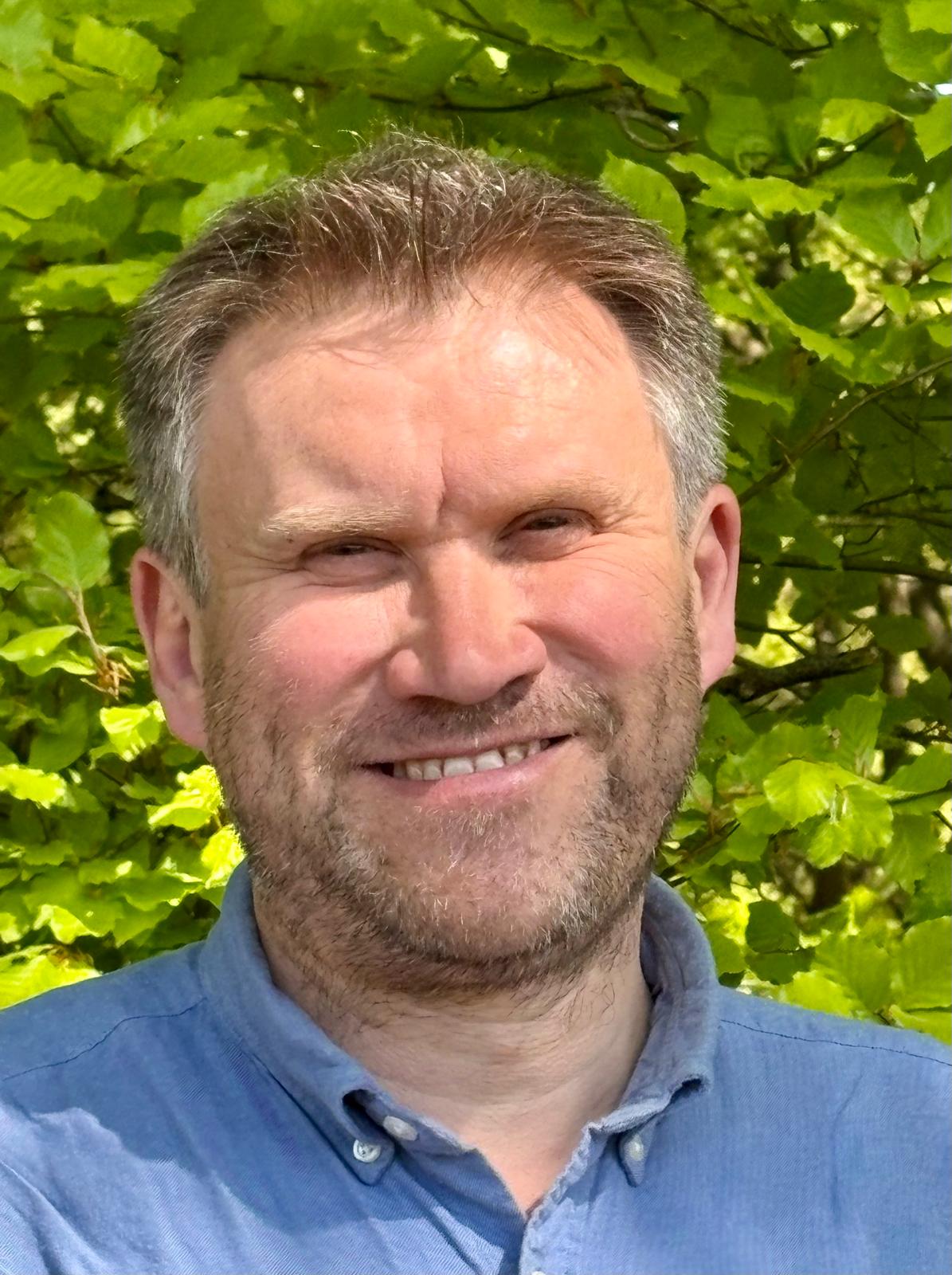 A headshot of Tom Ovenstone, wearing a blue shirt and smiling at the camera