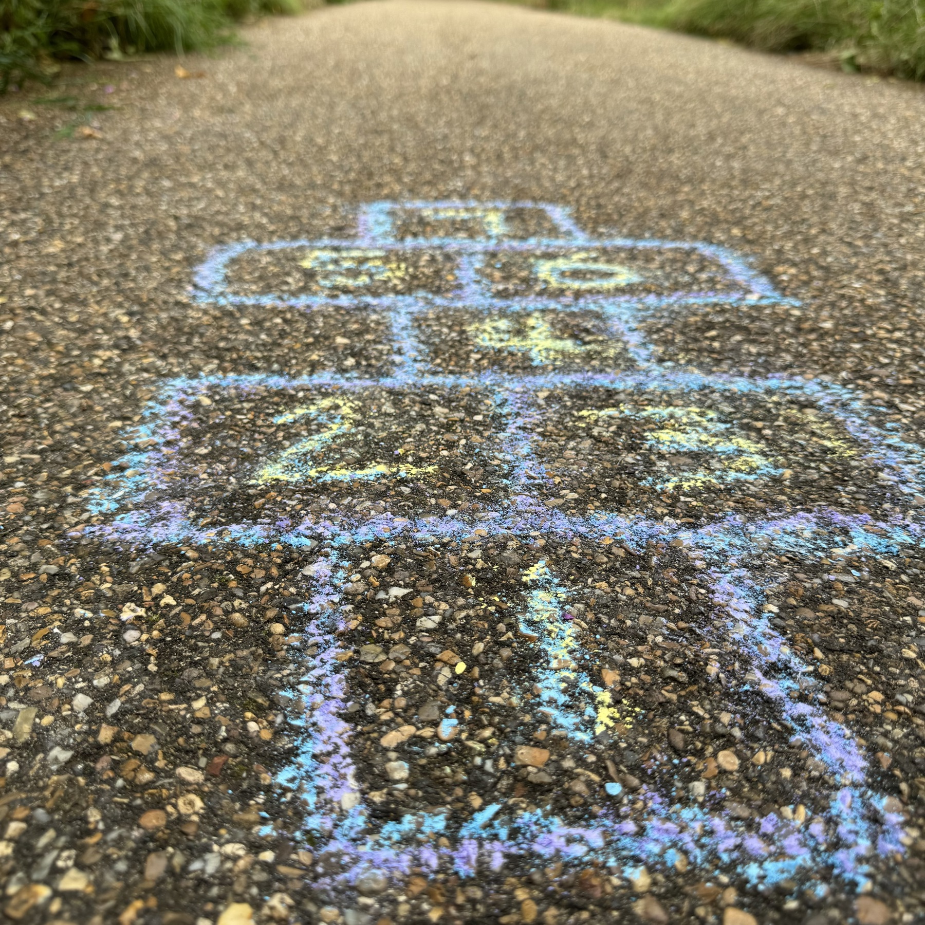 A pebble path that has squares drawn on it to make up a hopscotch ladder. It's drawn in blue chalk and purple chalk, with a number in each block in yellow chalk.