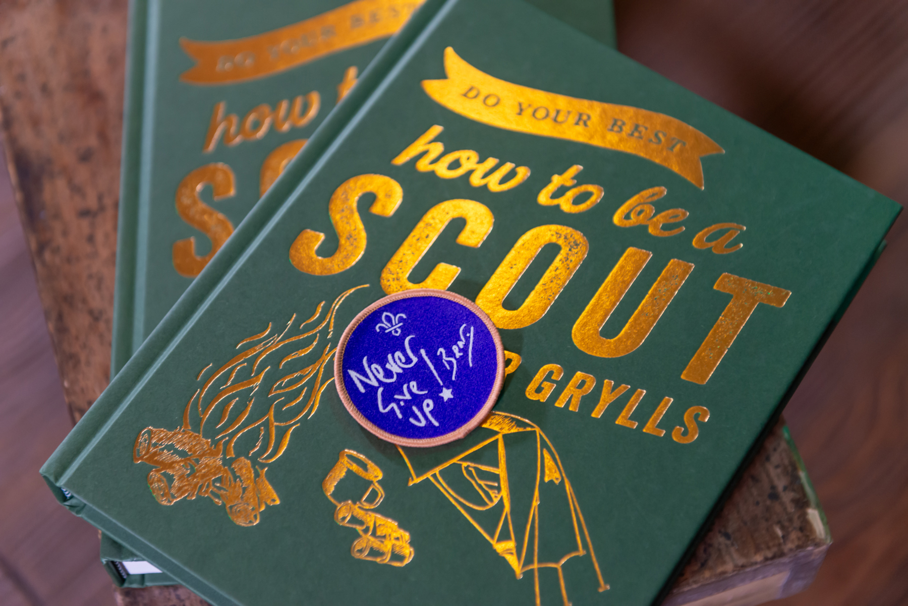 The image shows two copies of Chief Scout Bear Grylls' new book, Do Your Best: How to be a Scout, with a purple 'Never Give Up' badge on top of the cover. The books are laid on a wooden surface with one book on top of the other.