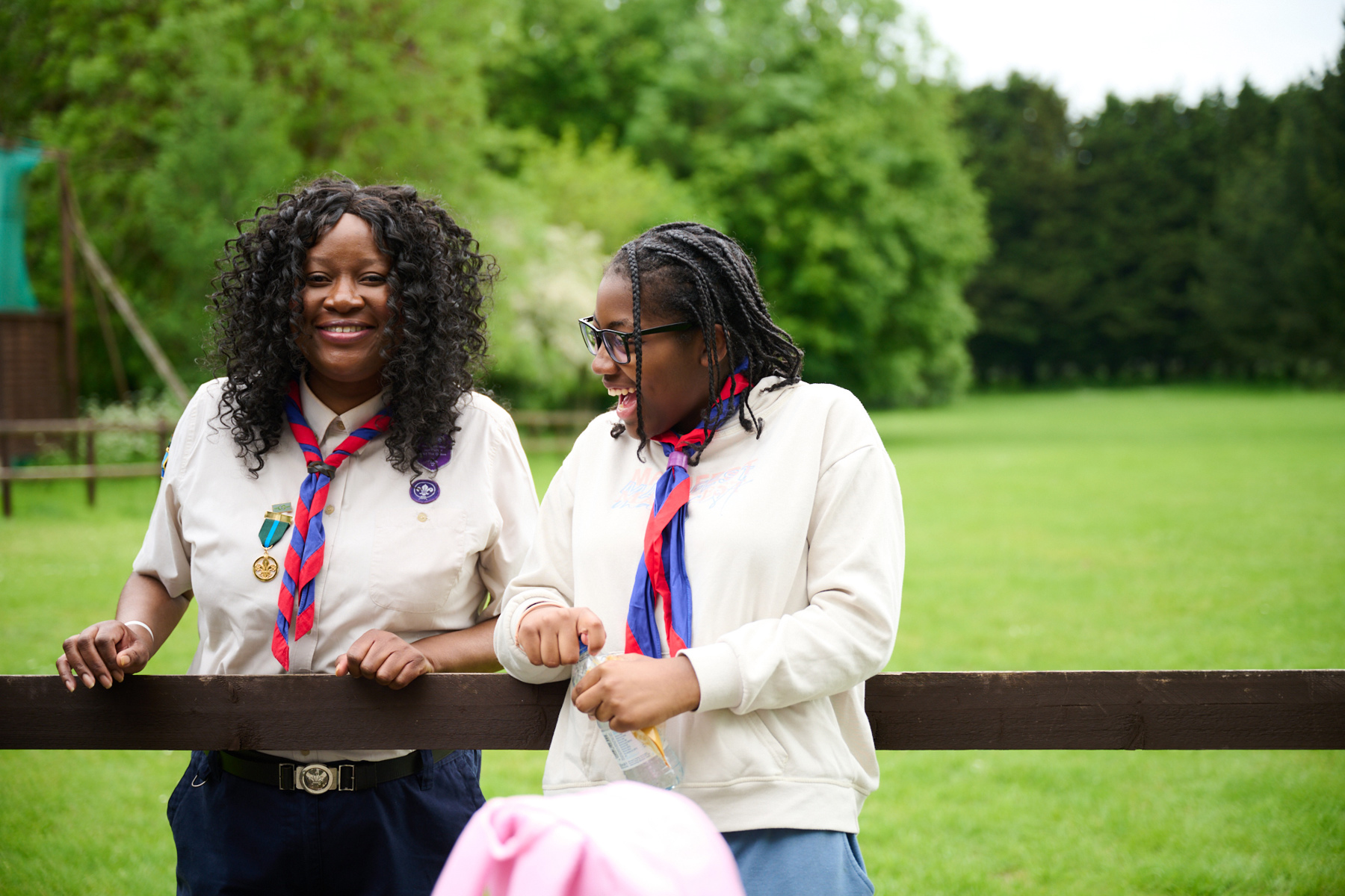 Two Black Scout volunteers are stood on grass. The volunteer on the left is behind a brown fence, while the volunteer on the right is in front of the fence and leaning on it. The volunteer on the left is smiling and looking at the camera, but the one on the right is laughing and looking to the left. 