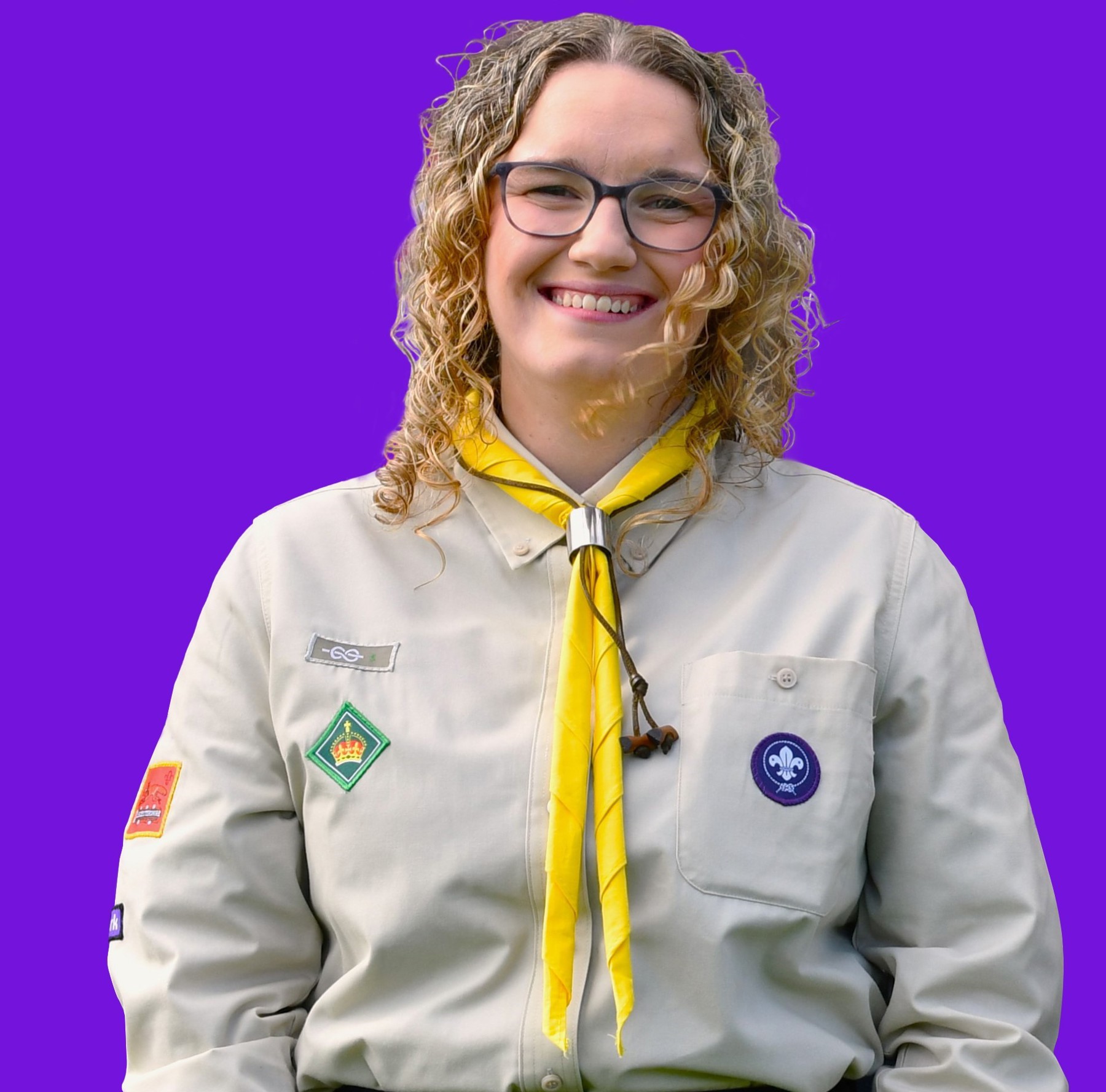 This image shows a woman with curly blonde hair wearing glasses and smiling at the camera in front of a plain purple background. She's wearing a Scouts uniform shirt and a yellow necker.