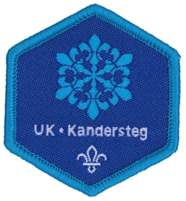 The image shows the blue UK - Kandersteg badge. It's navy blue with a lighter blue border, a white fleur-de-lis sitting at the bottom with the text 'UK - Kandersteg' just above. Just above that is the Kandersteg International Scout Centre logo in light blue. 