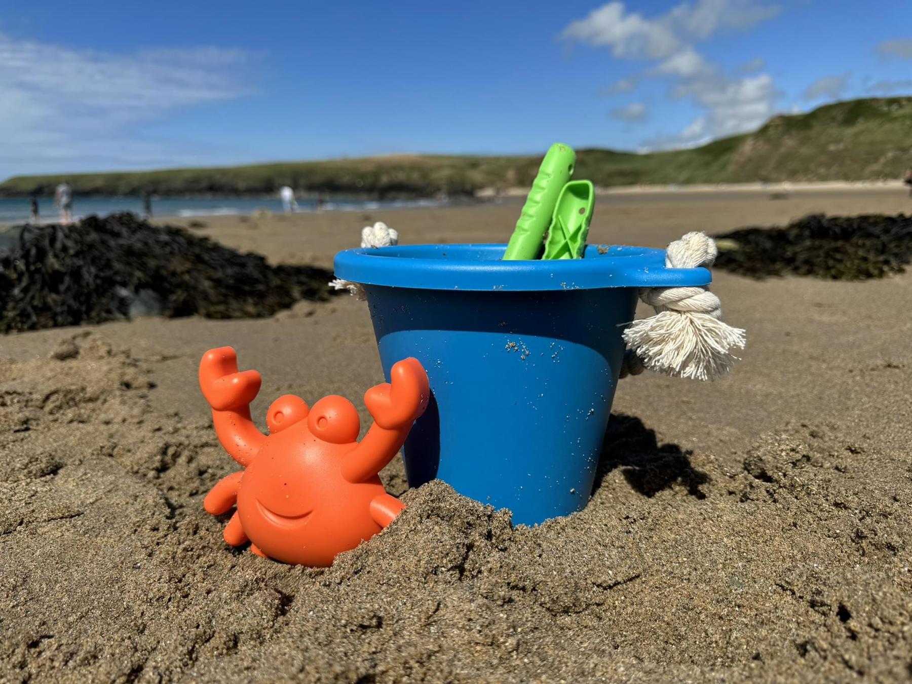 A blue bucket on a beach. It's placed int the sand with two green spades in it. An orange crab sand shaper is resting on the bucket. 