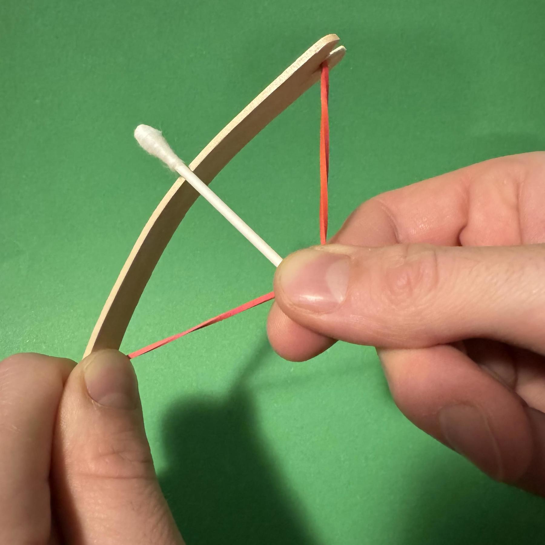 A mini bow and arrow on a green background. A cotton bud is the arrow and a red elastic band and lollipop stick make the bow. It's on a green background and a hand is holding the cotton bud arrow on the elastic band, then pulling them back to fire it.